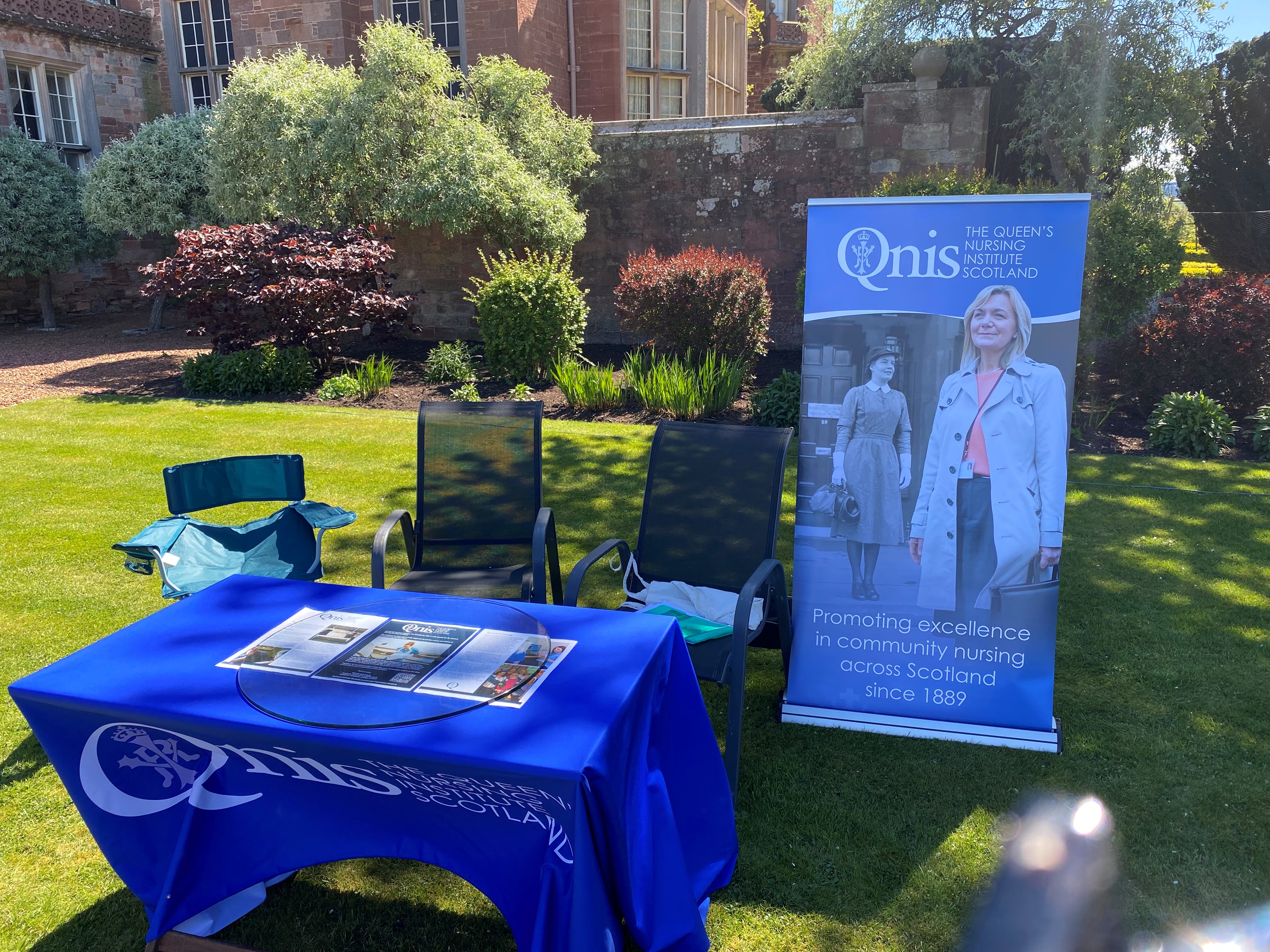 A table with a blue table cloth is next to a banner advertising the QNIS. It is set up in a garden on a sunny day.
