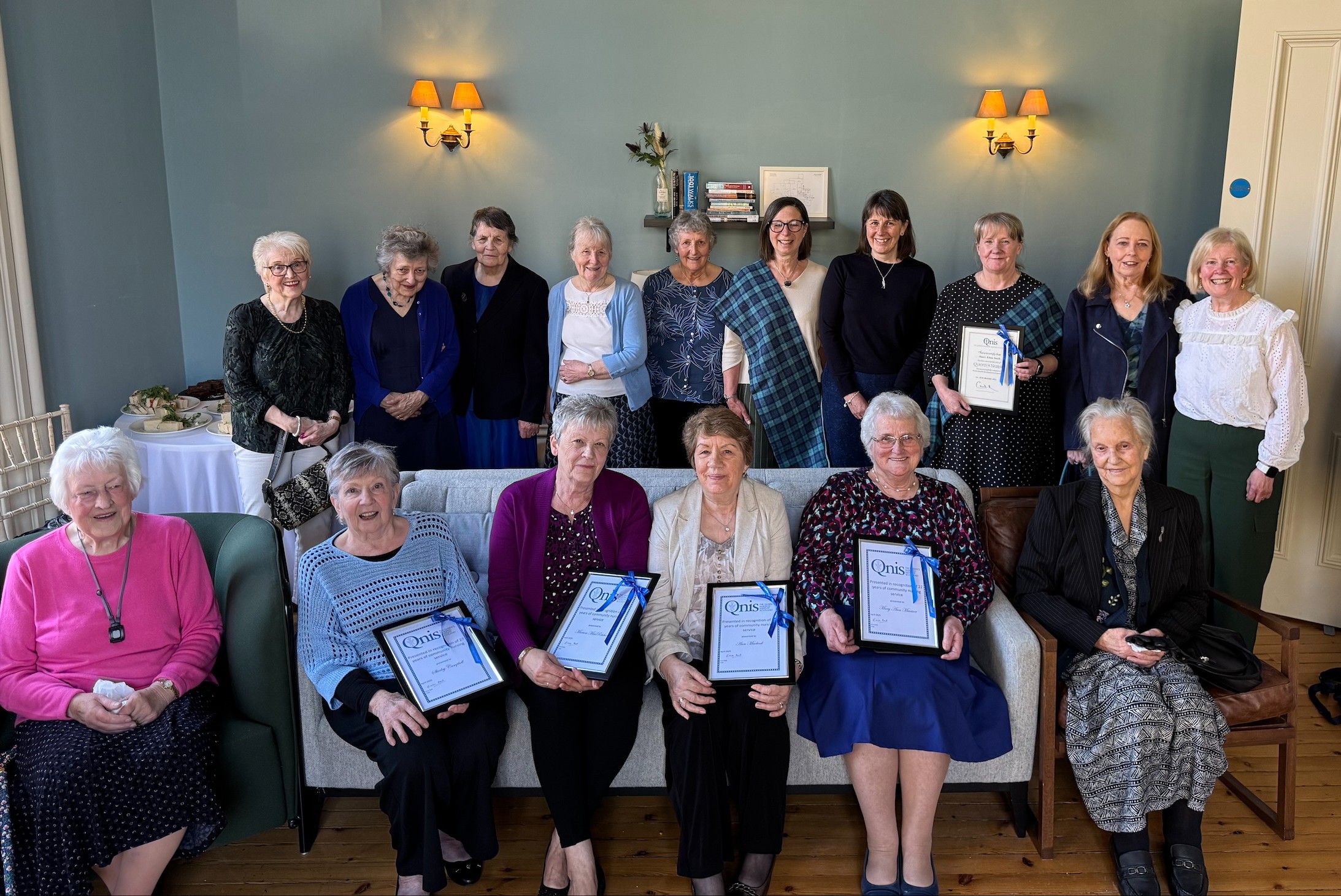 A colour image of a group of older adults smiling and seated and standing together. Five of the group hold framed certificates. There is a pale blue wall behind the group with a light at either side.