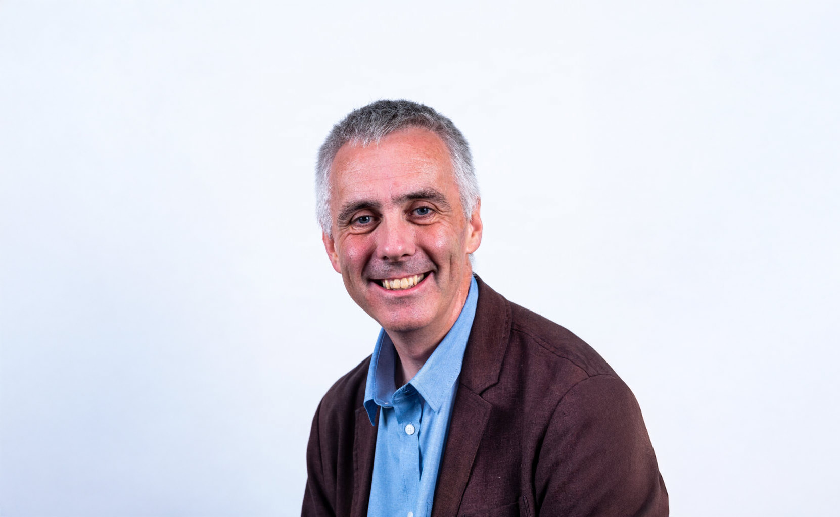 A person with short grey hair smiles at the camera. They are wearing a light blue button-up shirt under a brown blazer, seated against a plain light background.