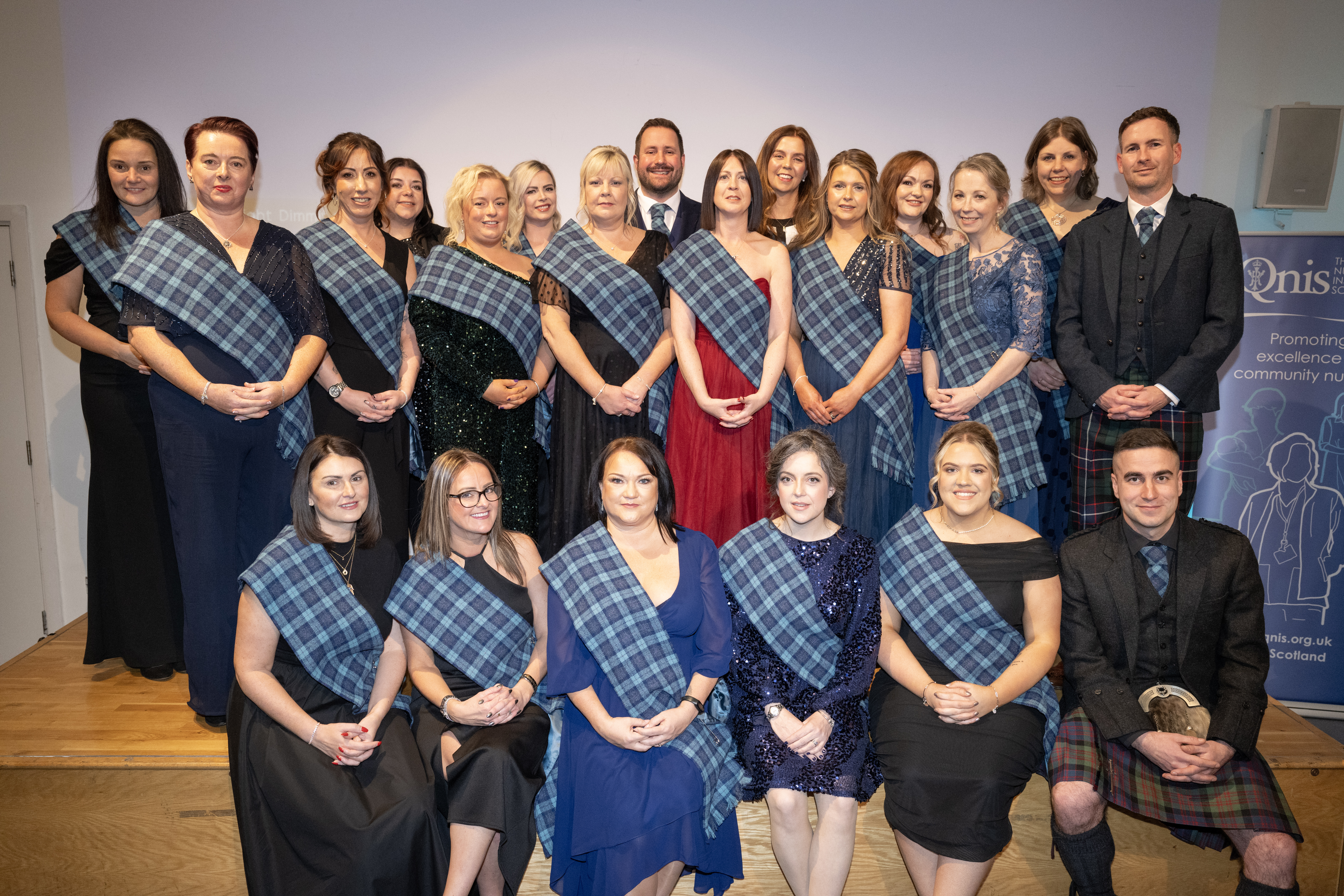 A group of 21 people pose for a group photo indoors. They are all wearing formal attire, long dresses in a range of colours and three people in Scottish dress with kilts. Apart from these three, all wear a blue tartan sash diagonally across their torso. They are smiling and looking at the camera. The group is arranged with two standing rows and six sitting at the front. They are standing or sitting on a pale wooden stage.