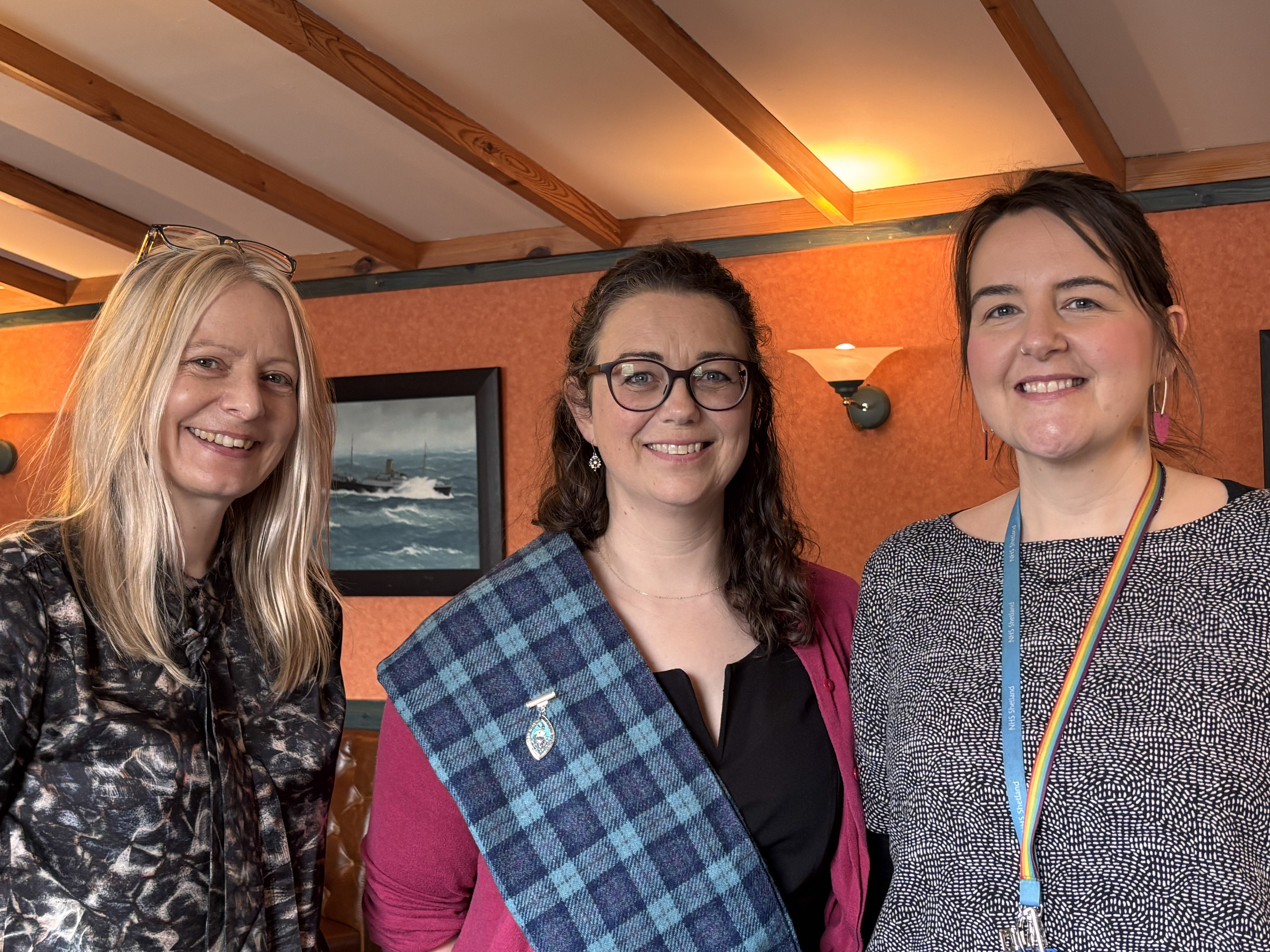 Three people stand side by side indoors, smiling at the camera. The room has warm lighting, wooden ceiling beams, and an orange-toned wall with a framed painting of a boat behind them. One person wears glasses and a blue tartan sash, another has long blonde hair and a patterned top, and the third wears a lanyard over a dark patterned dress.