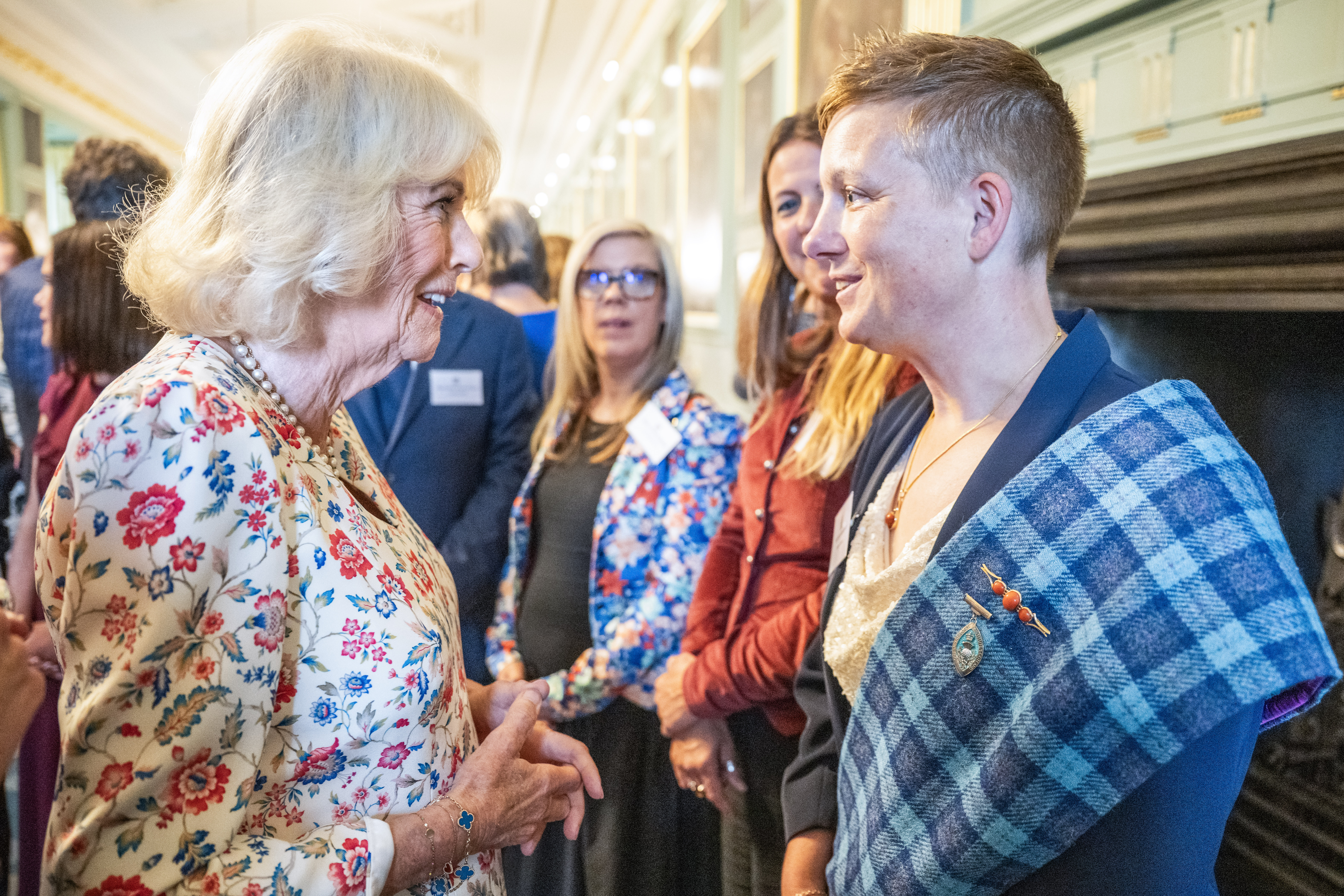 Two people are in deep conversation whilst others look on. One of the people is wearing a tartan sash. The other person is wearing a floral dress.