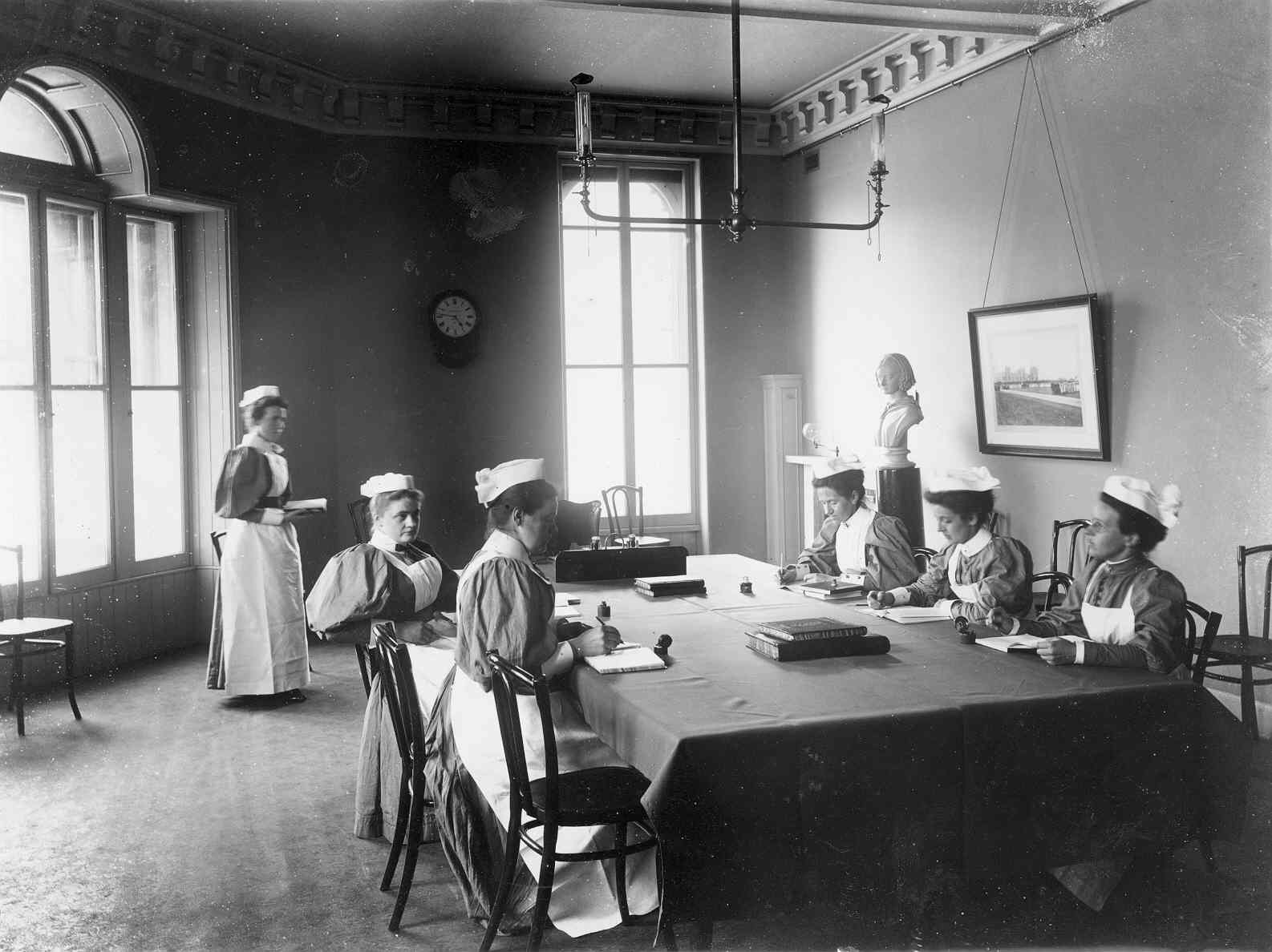 The move to Castle Terrace   A black and white image of six people in a large room. Five people sit at a large table in the centre of the space, one person stands. They all wear traditional nursing uniforms, long dress, light-coloured apron and white bonnet-style hat.
