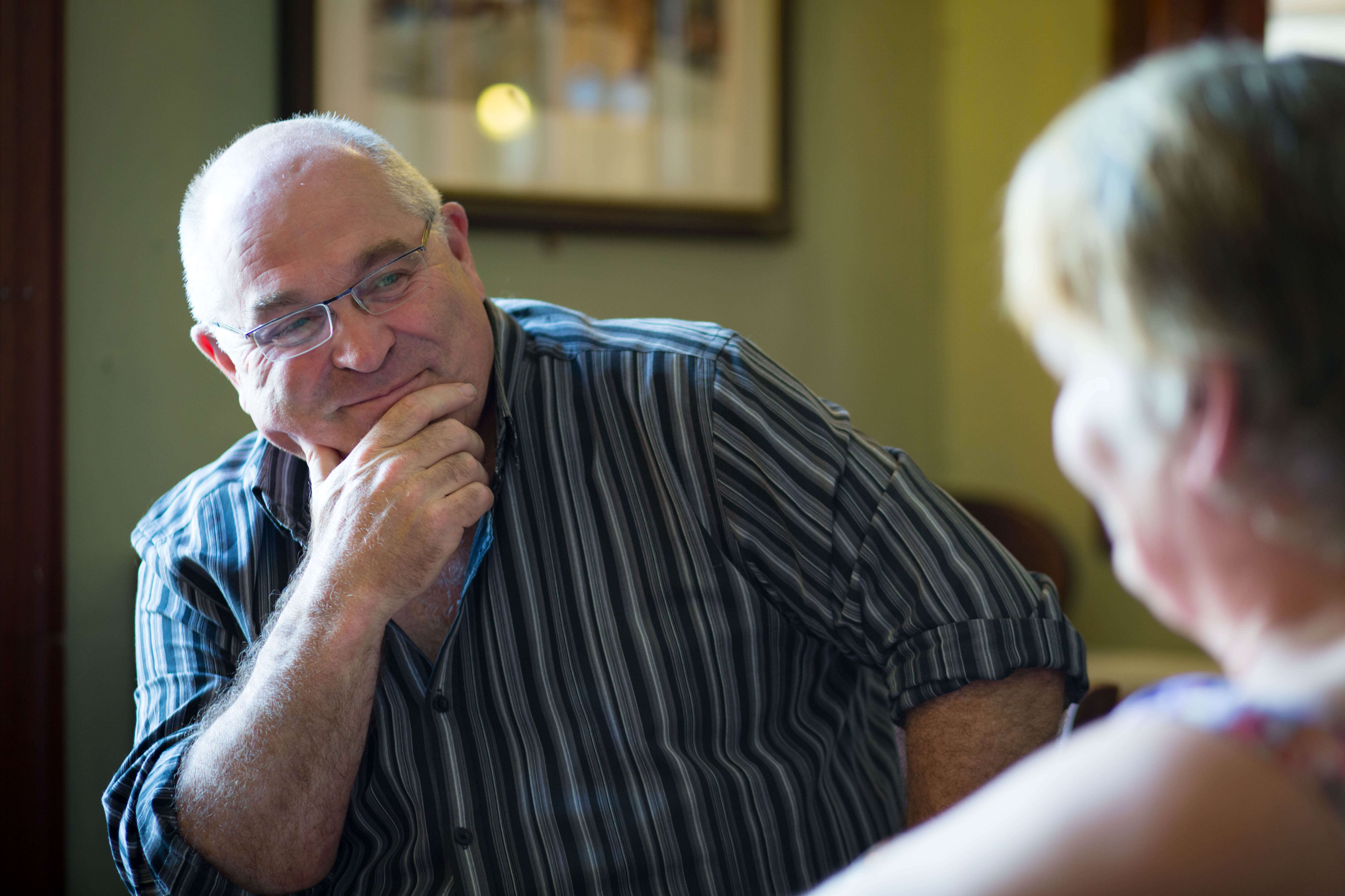 A person wearing glasses and a striped shirt sits indoors, smiling thoughtfully with a hand resting on their chin, engaged in conversation with another person who is out of focus in the foreground. The background is softly lit, featuring a framed picture on the wall.