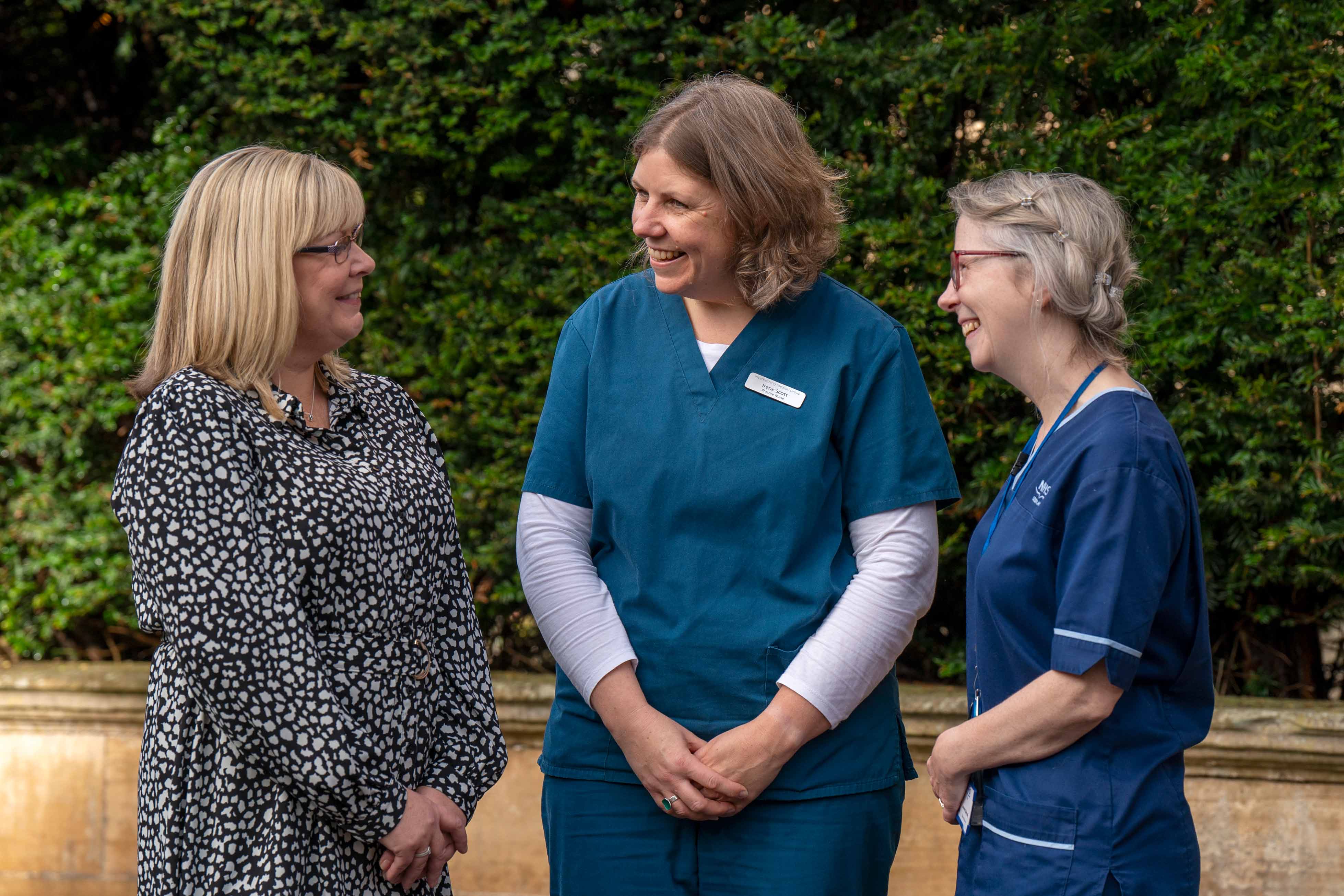 Three people standing outdoors, smiling and talking together. One person wears a patterned black and white dresses, while the other two wear medical uniforms, one in teal and the other in navy blue. They are standing in front of greenery.