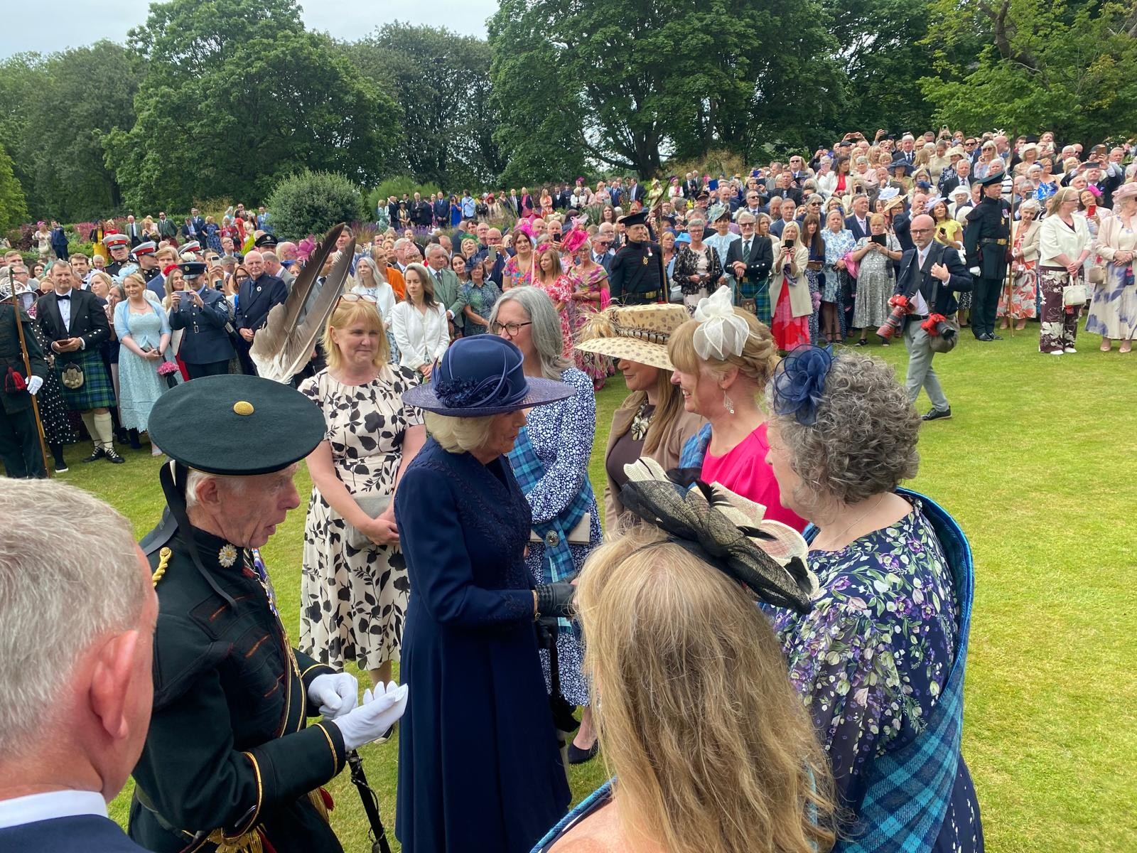A group of people in formal attire stand together in a garden. Three of the people are wearing tartan sashes.. They are chatting and smiling.