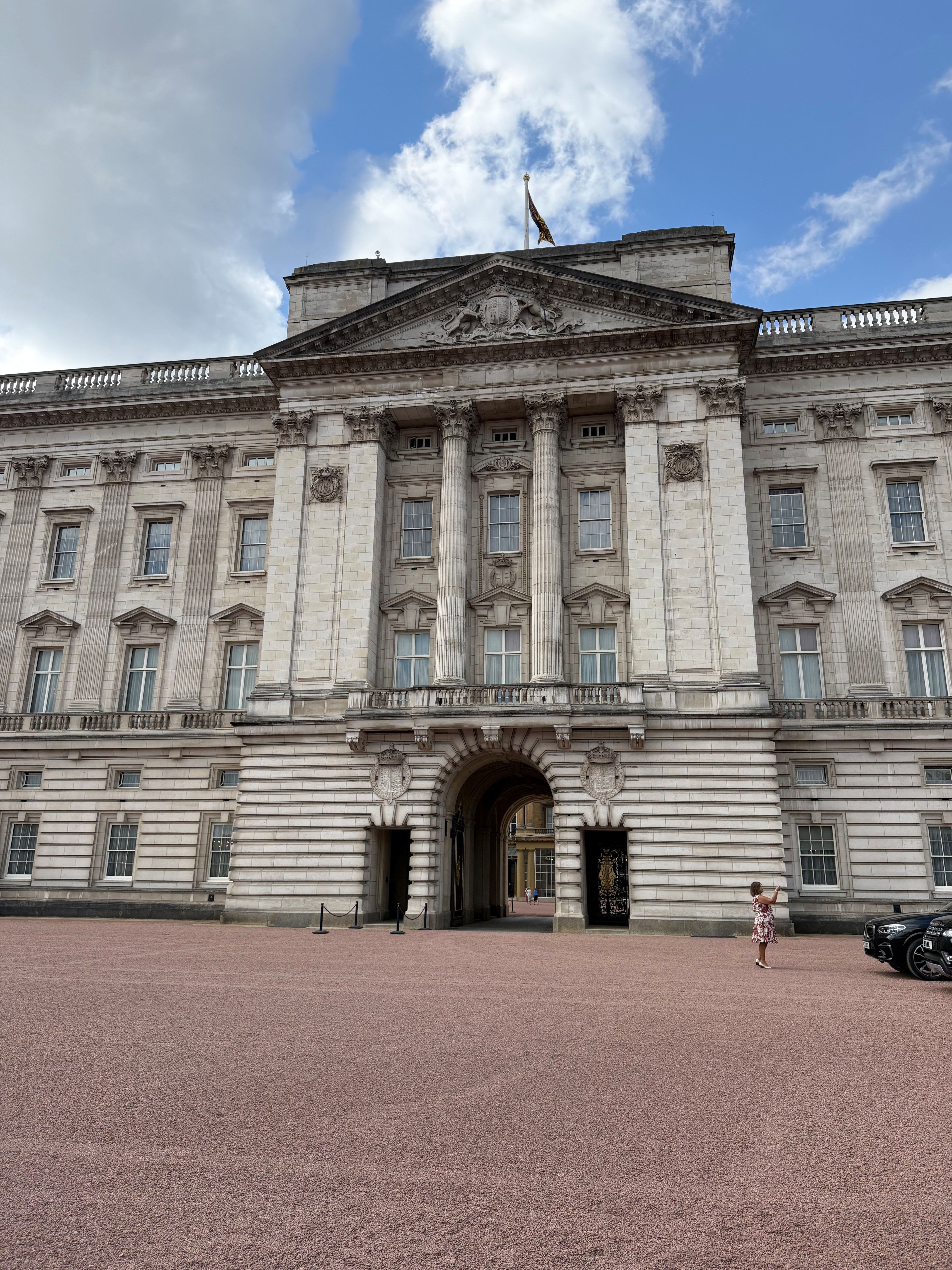 Front view of a large, ornate stone palace with tall columns and an arched central entrance, set against a partly cloudy sky. A wide gravel courtyard stretches across the foreground, where a single person stands off to the right taking a photo.