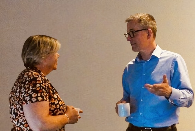 Two people standing and having a conversation indoors. One person is wearing a floral-patterned top, and the other is wearing glasses and a light blue shirt while holding a white mug. They are facing each other and appear engaged in friendly discussion against a neutral background.