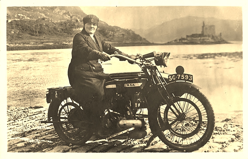 A sepia-toned photograph of a person sitting on a vintage BSA motorcycle, parked on a cobblestone surface beside a body of water. The bike registration is SG-7593. The person is wearing a long coat and a cap and is looking toward the camera with a neutral expression. In the background, across the water, is a mountainous landscape with a structure resembling a castle or ruins on an island. 