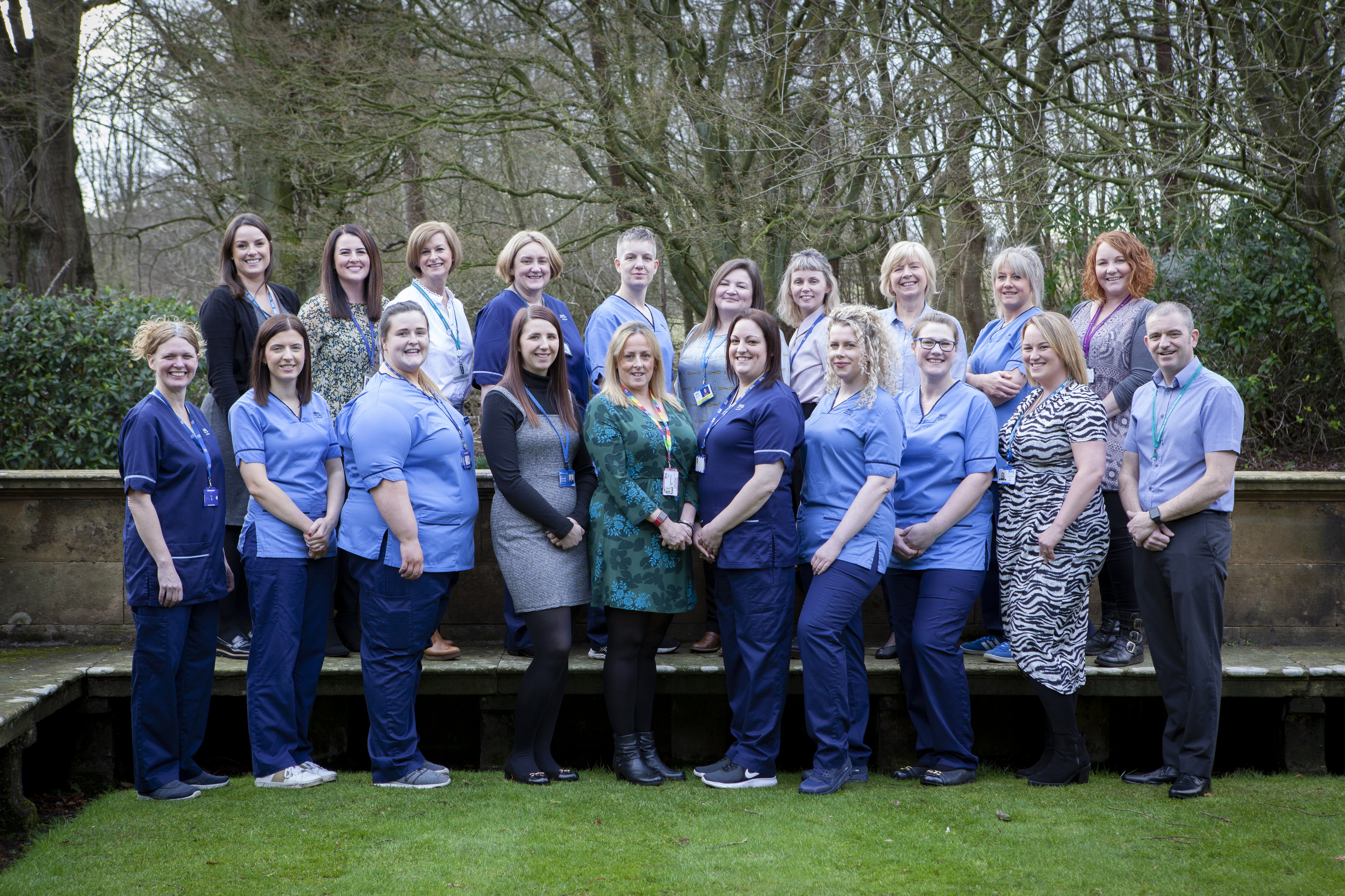 A group of 20 people pose for a group photo outdoors in front of a stone ledge and a backdrop of trees. Most individuals are wearing professional uniforms, primarily in light blue and navy, while others wear business or casual attire. The group is arranged in two rows, with some standing on a raised stone platform. The setting appears to be a park or garden area during winter or early spring, with leafless trees in the background and green grass underfoot. All individuals are smiling and facing the camera.