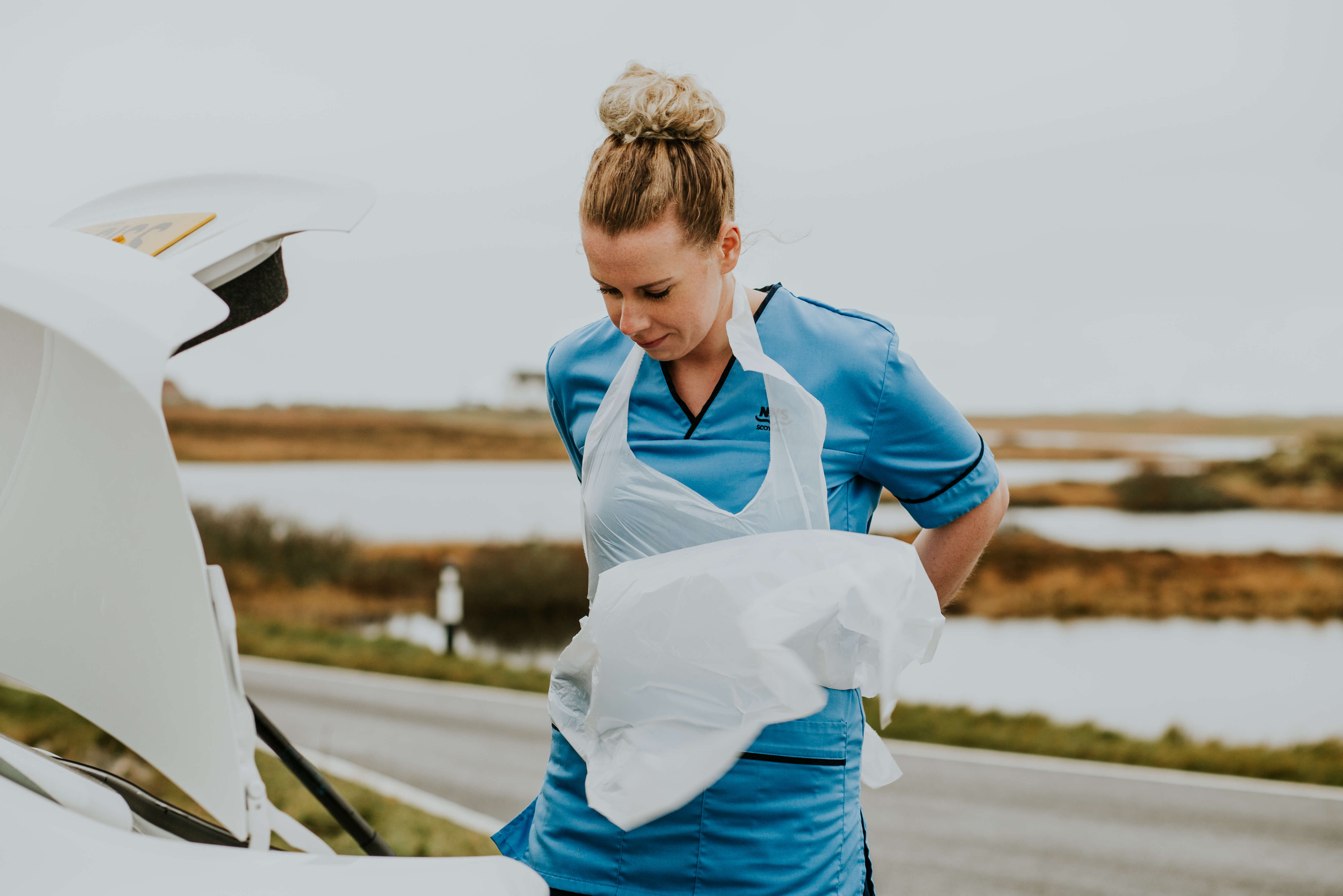 A nurse wearing a blue uniform and a white plastic apron stands beside an open car boot on a rural roadside, with water and grass visible in the background.