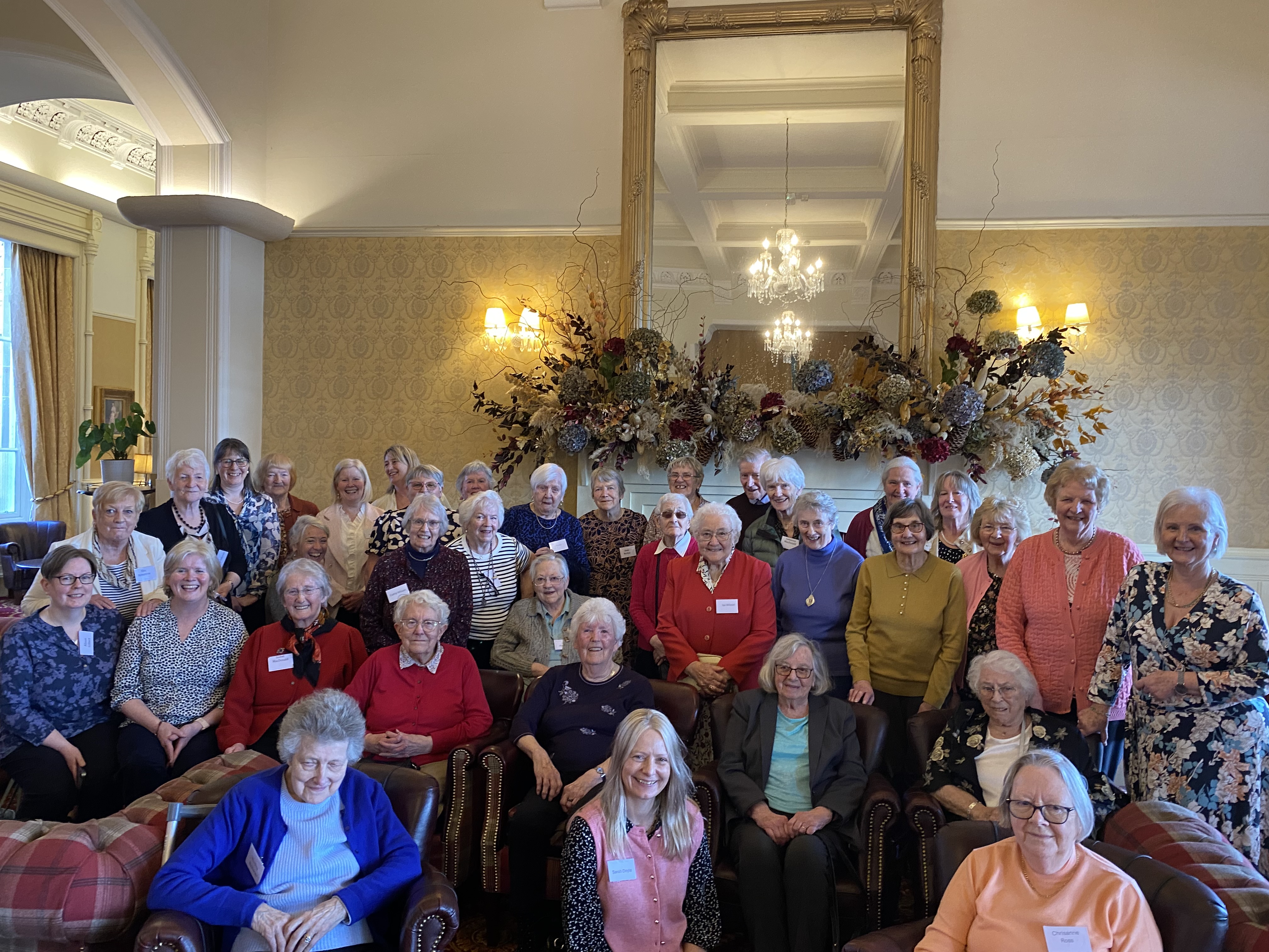 A large group of older adults pose together in a bright, elegant room with patterned wallpaper, a tall mirror, and a decorative floral arrangement above a fireplace. Some are seated in armchairs at the front while others stand behind them. Many wear name tags and smile toward the camera, suggesting a social gathering or community event.