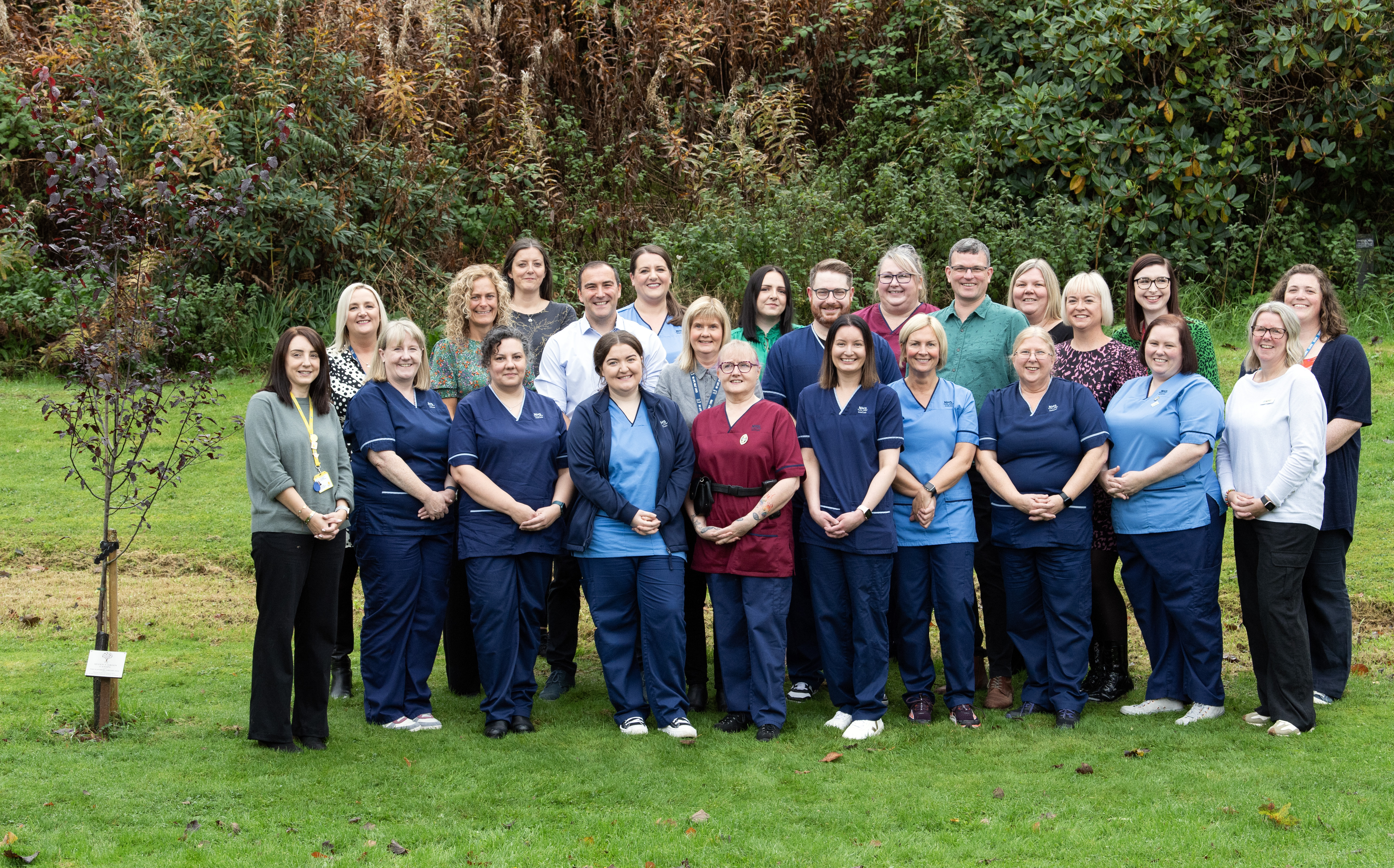 A group of 24 people pose for a group photo outdoors. Most individuals are wearing professional uniforms, primarily in maroon, light blue and navy, while others wear business or casual attire. The group is arranged in three rows. The setting appears to be a park or garden area, with trees in the background and green grass underfoot. To the left of the people a newly planted tree with a white label near the base of its trunk. All individuals are smiling and facing the camera.