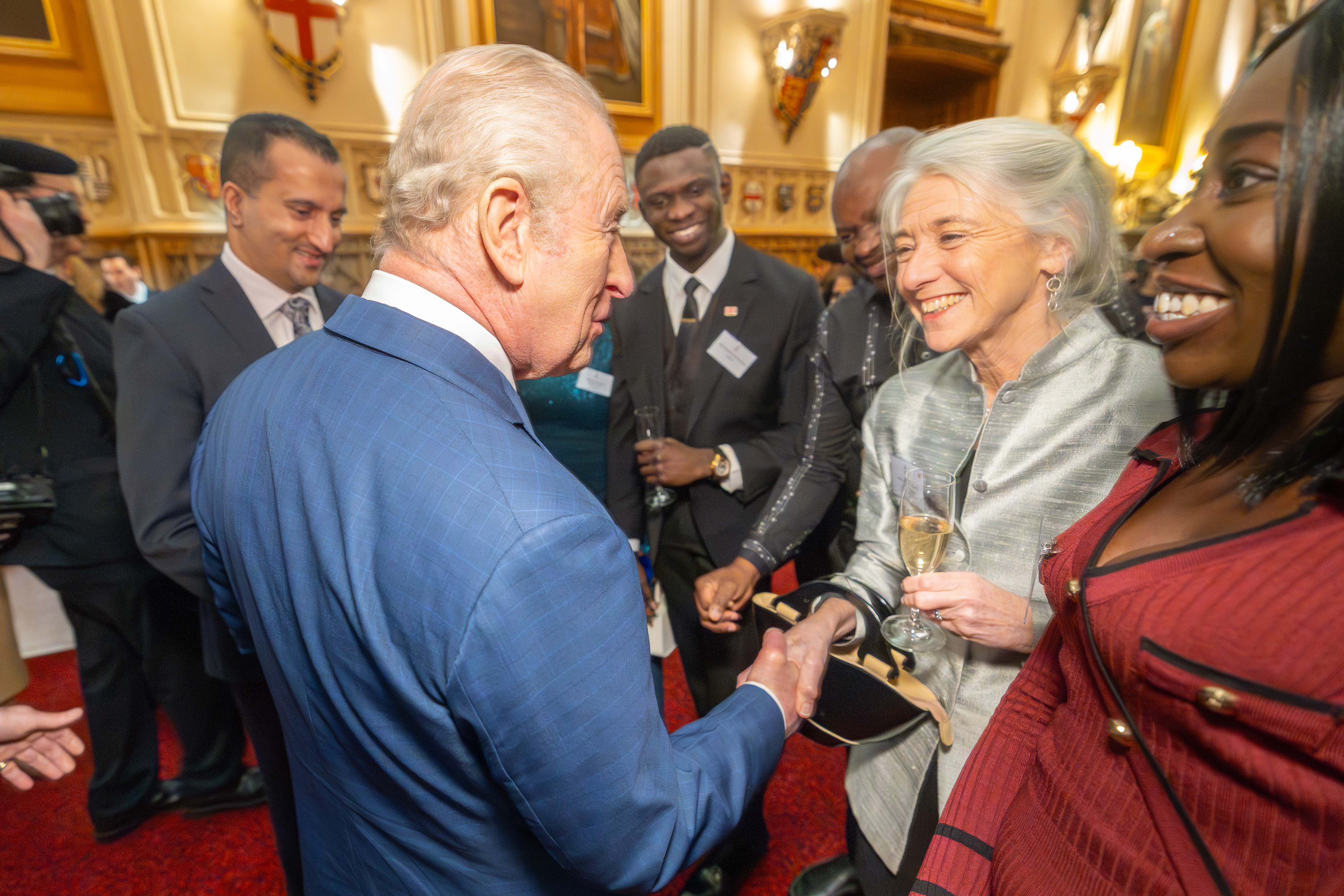 Two people with white hair greet each other in a formal setting. One wears a blue jacket, the other a sliver jacket. There are four smiling people looking on.
