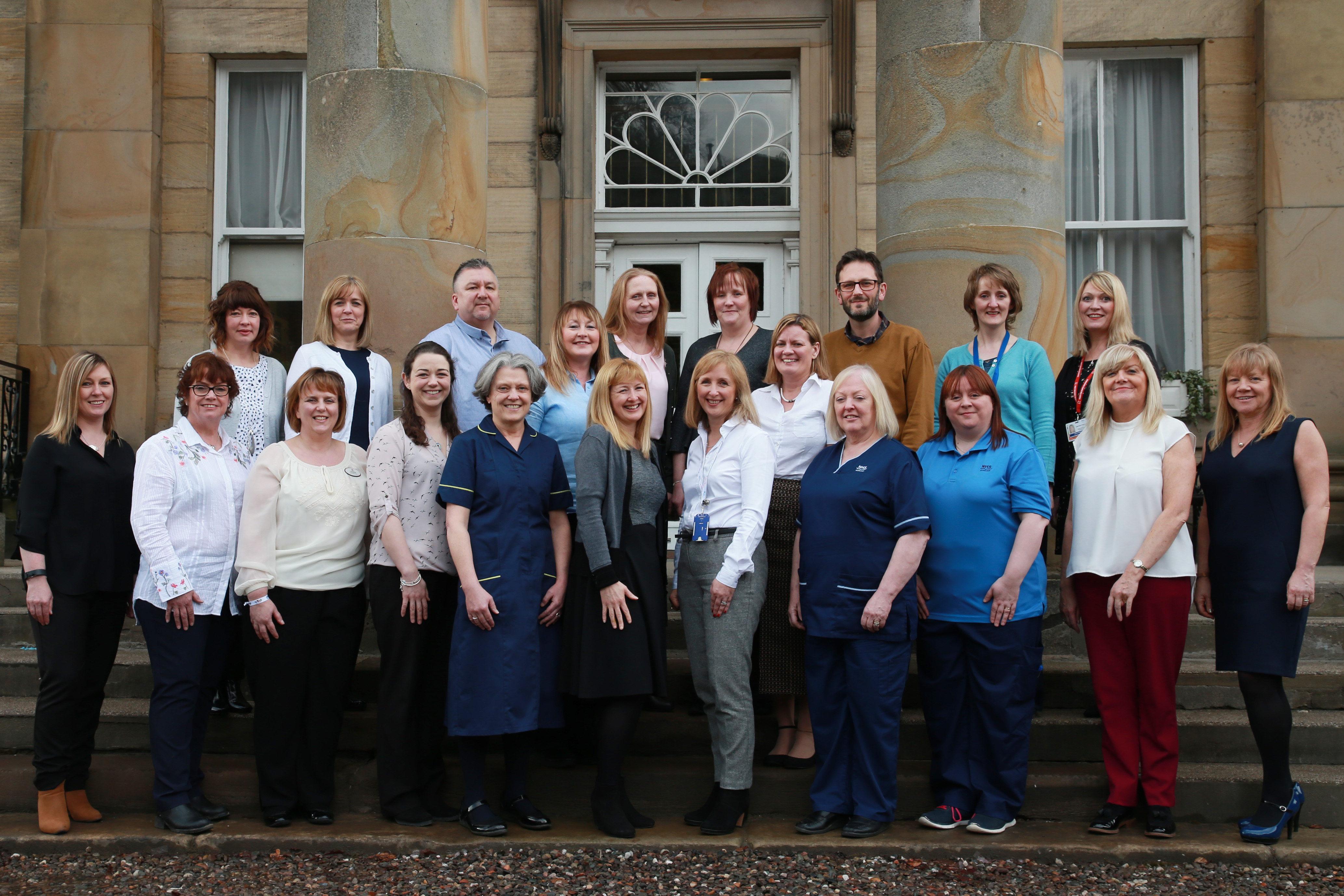 A group of 21 adults posing for a group photo in front of a large stone building with two tall columns and a decorative window above the door. The group is arranged in two rows, with some standing on the steps and others on the ground. They are dressed in a mix of professional attire, including office clothing and uniforms, suggesting a workplace setting. The expressions are generally friendly, and the photo appears to have been taken during the day in an outdoor setting.