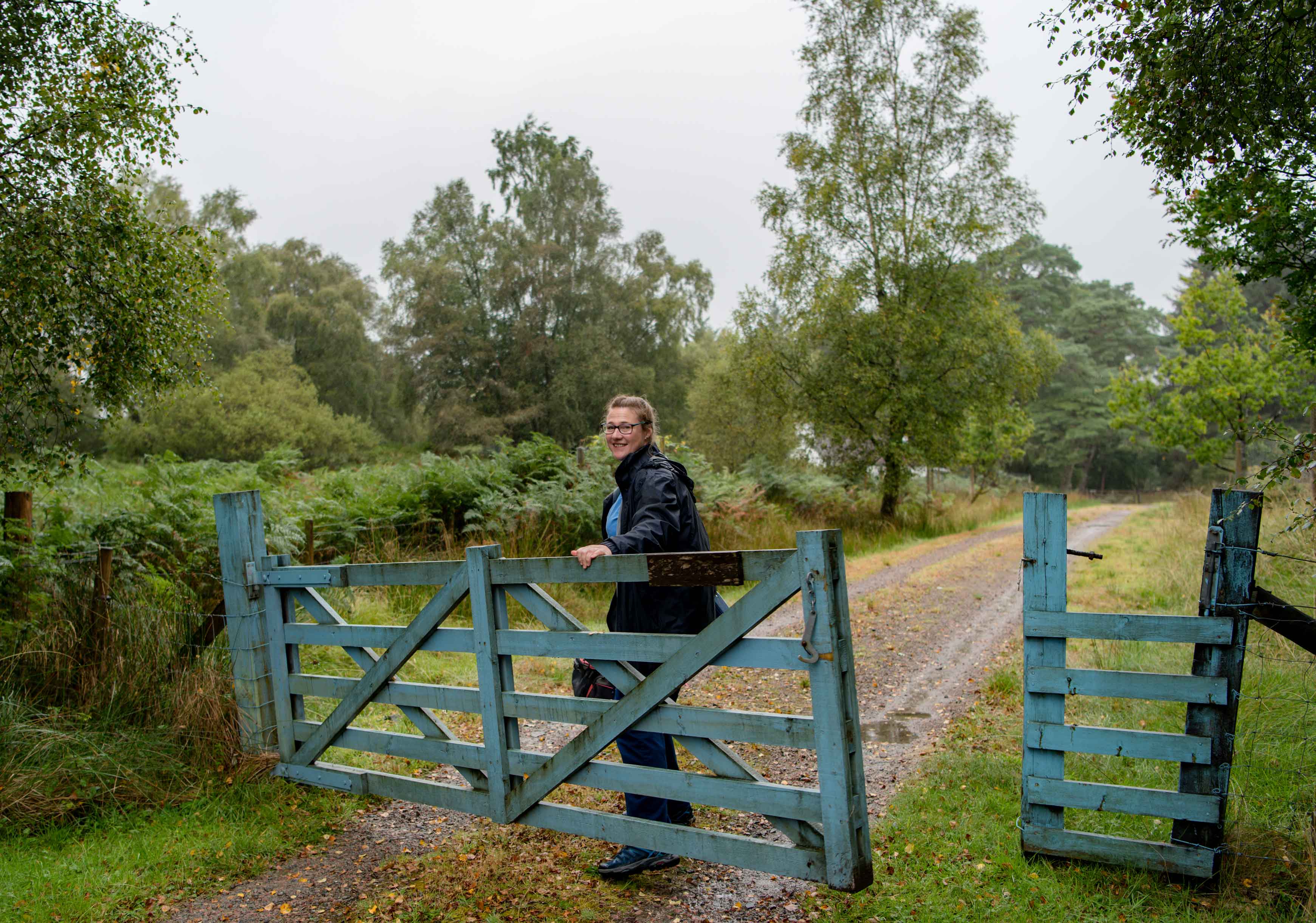 A person in a rain jacket smiles while holding open a blue wooden gate on a rural path surrounded by green trees and bushes. The sky is overcast, and the ground is wet, suggesting recent rain.