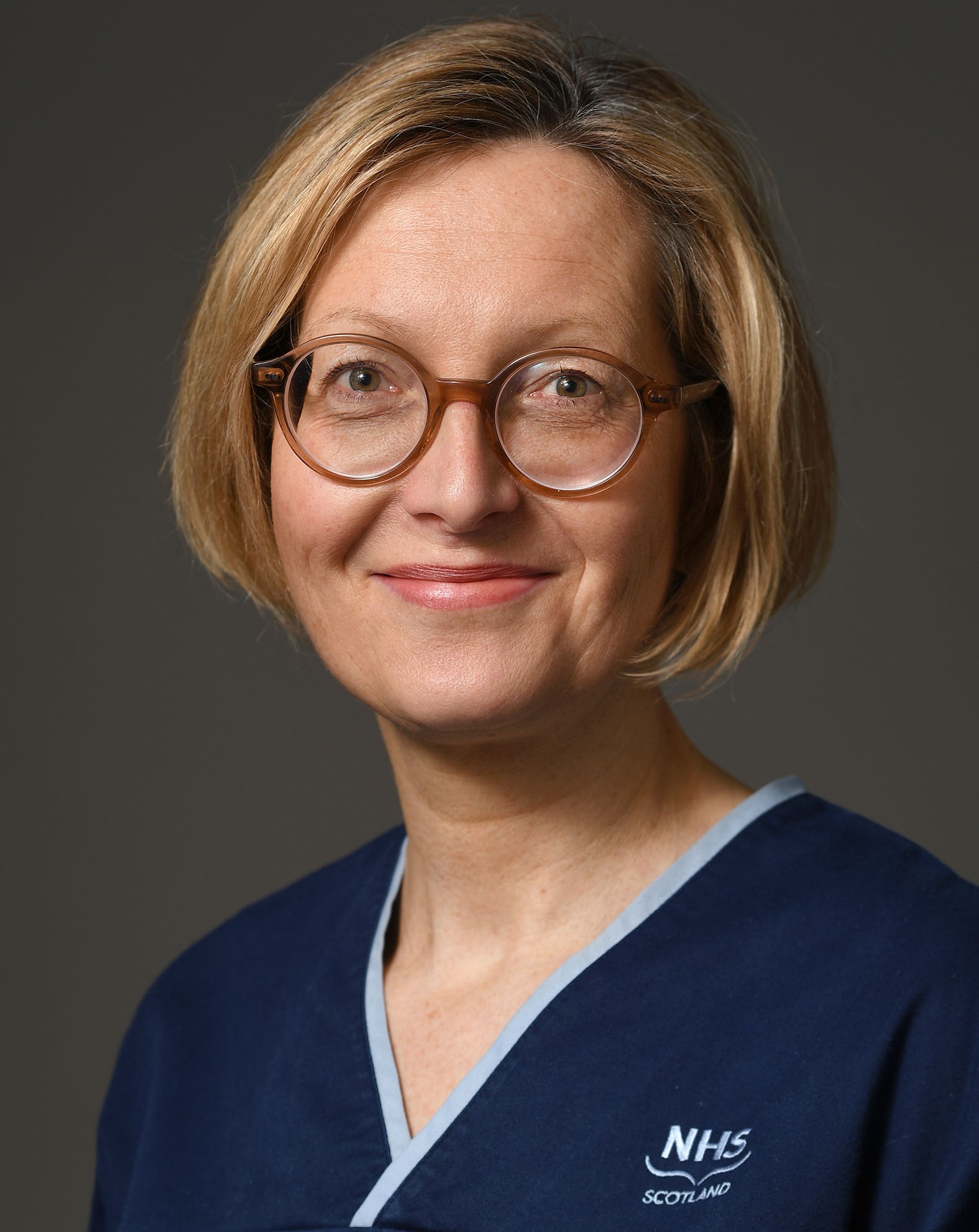 A person with short, light brown hair and round glasses is smiling slightly. They are wearing a navy blue uniform with light blue trim and an embroidered logo that reads 'NHS Scotland.' The background is plain and neutral.