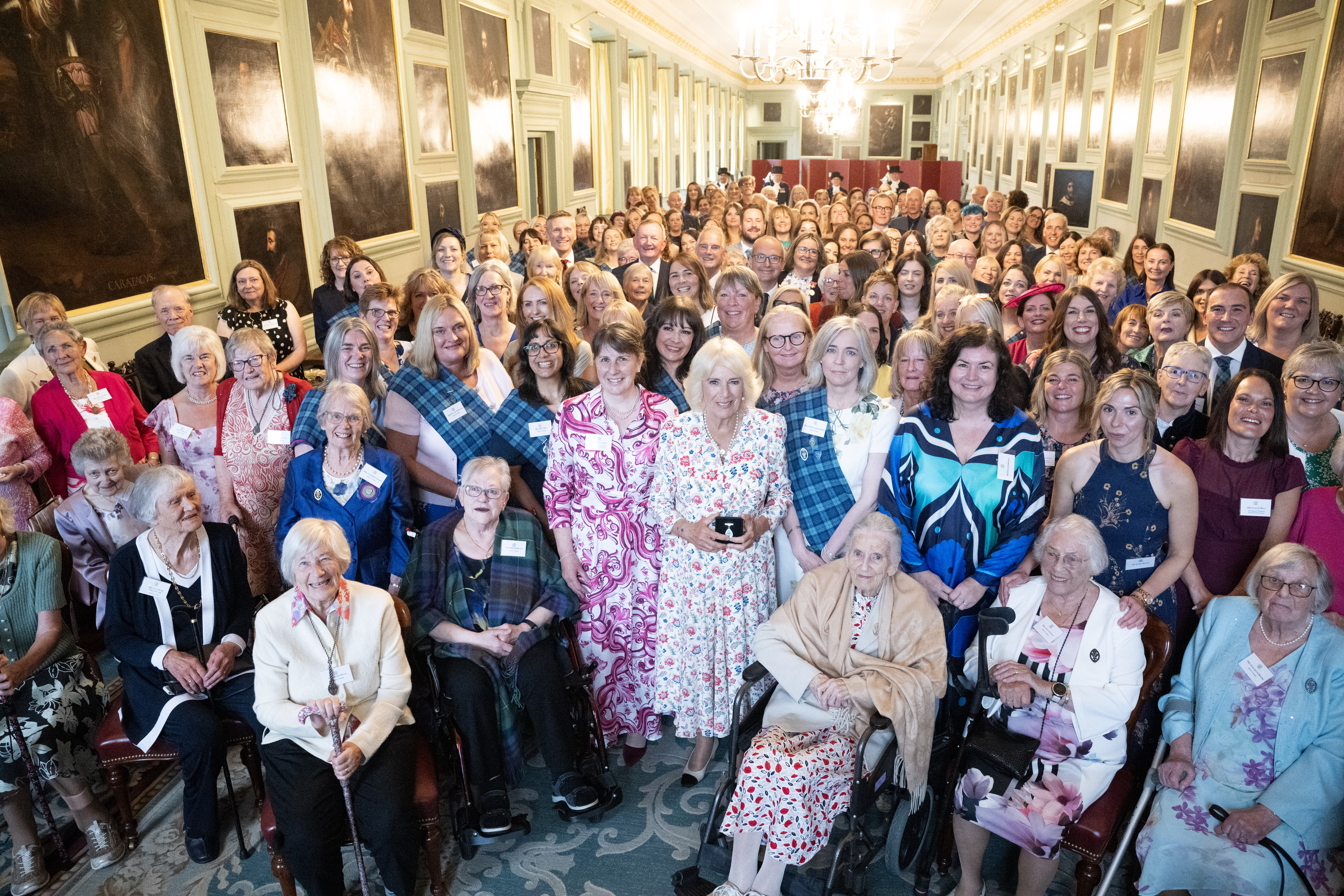 A group of people stand together smiling. They are in a long room with grand paintings on the wall. The person at the front holds a silver badge.