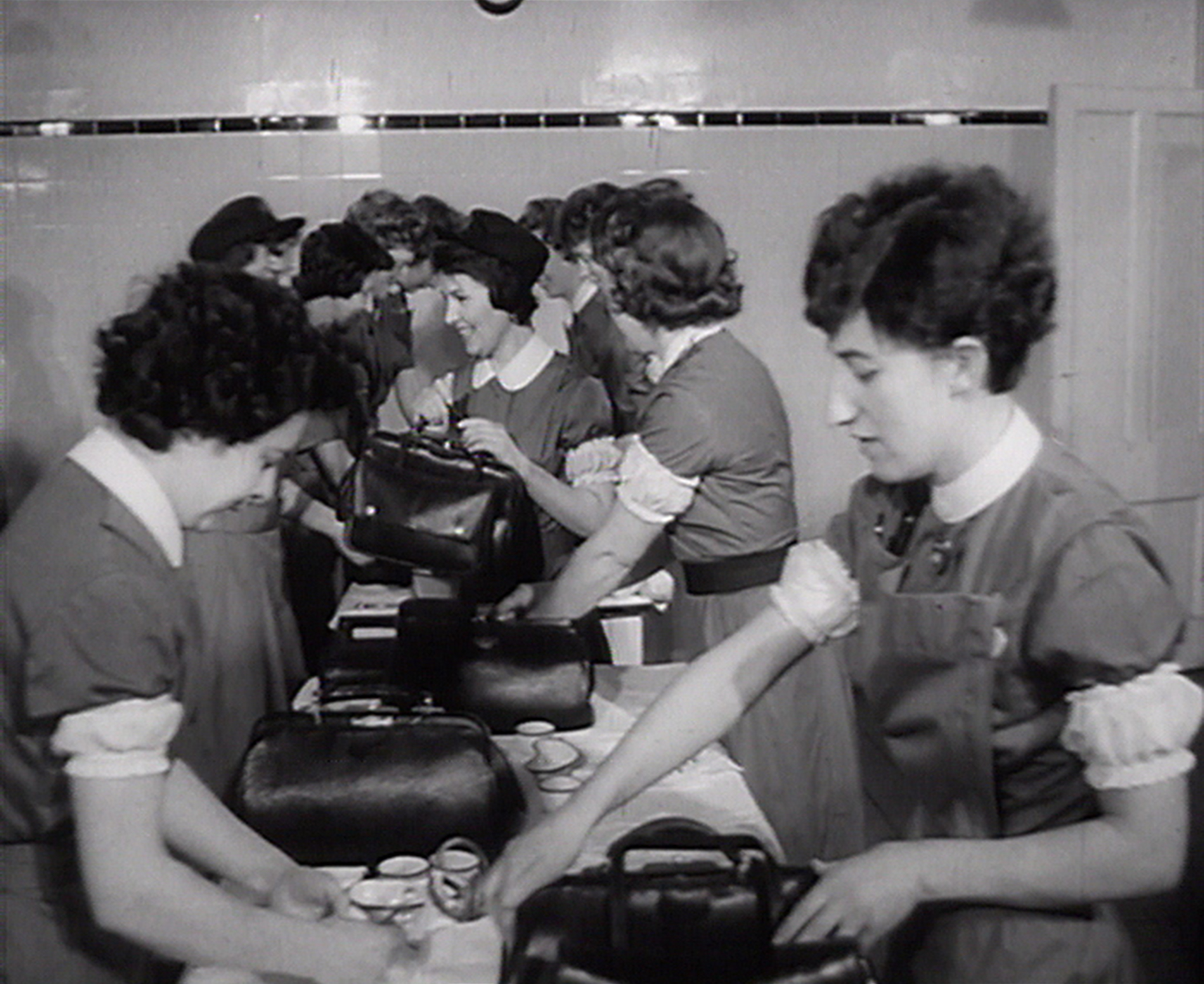 A black-and-white photo showing a group of people in uniform standing at a long table. They appear to be in a communal setting, possibly a cafeteria or break room. Each person has a large bag on the table in front of them, and they are interacting with items next to the bags. The individuals are engaged in conversation and activity, with some smiling. 