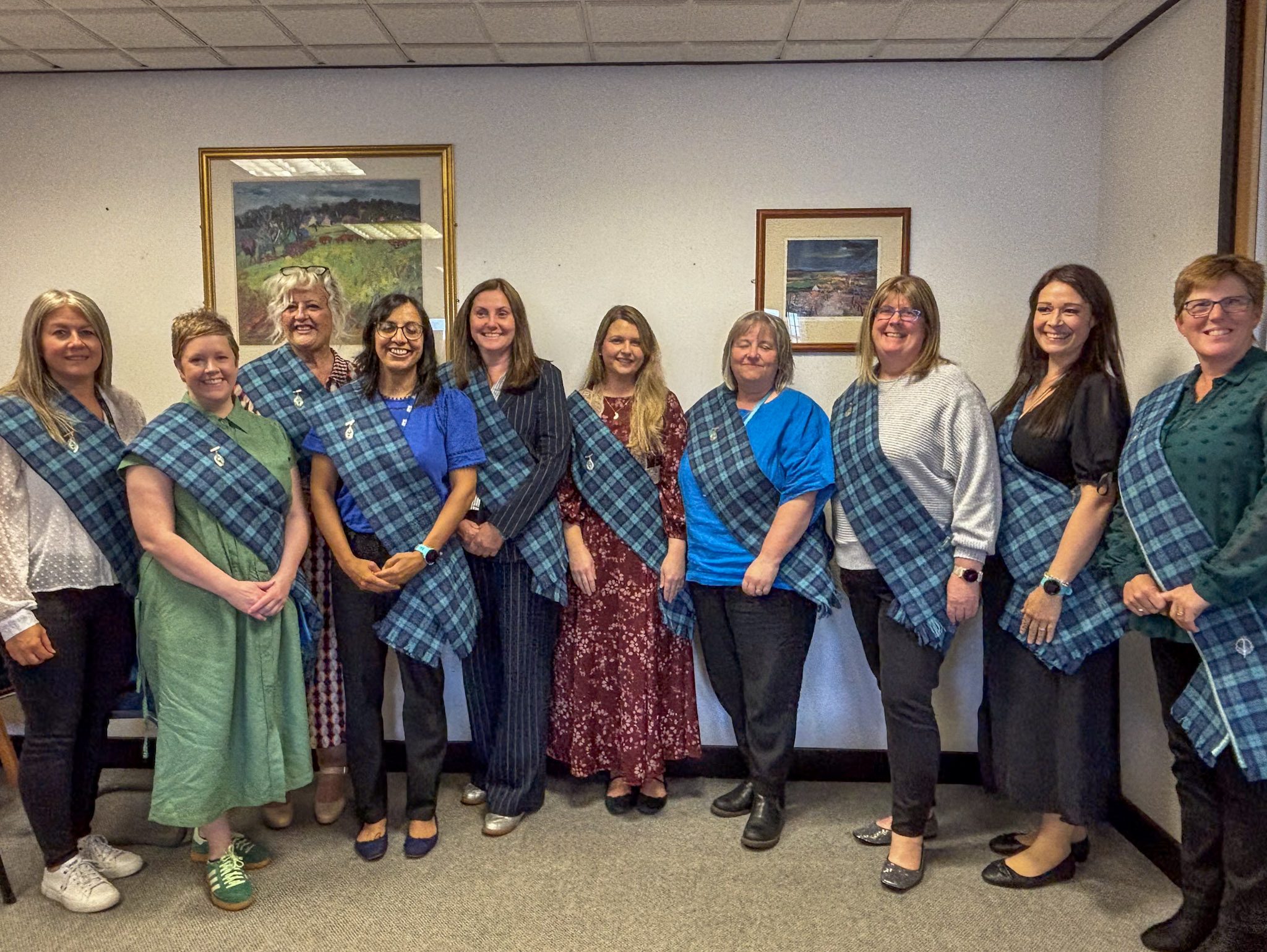 A group of ten people standing in a line indoors, smiling at the camera. Each person is wearing a blue and black tartan sash over their shoulder, secured with a circular pin. They are dressed in a mix of casual and smart-casual clothing. The background features a plain wall with two framed landscape paintings. The setting appears to be an office or meeting room with carpeted flooring and a tiled ceiling.
