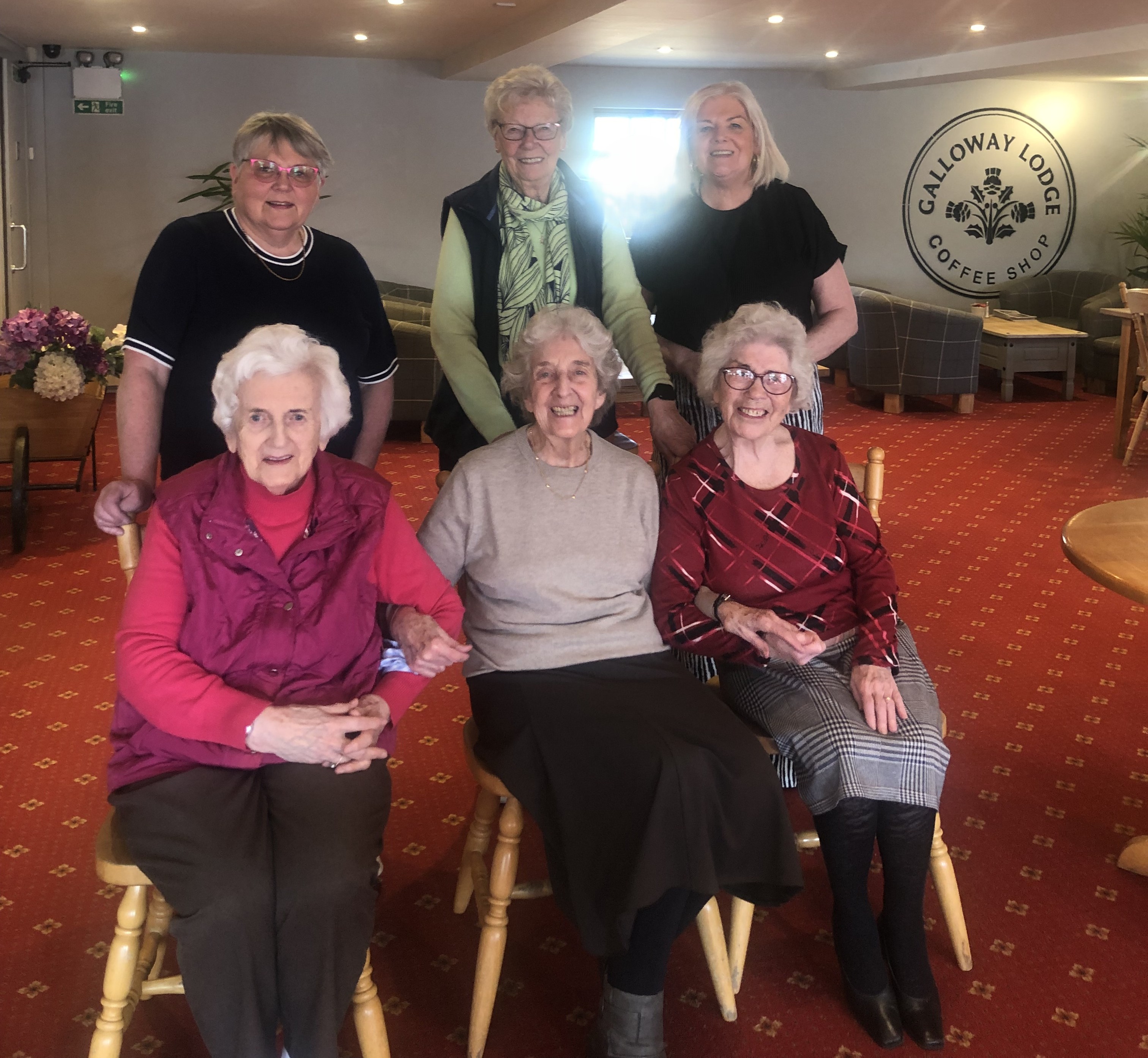A colour image of a group of six older adults smiling and seated and standing together. There is red carpet on the floor and a large circular sign saying Galloway Lodge coffee shop on the wall behind.