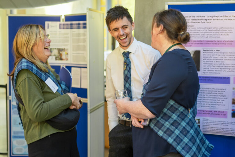 Three people are standing and laughing together in front of informational posters at what appears to be a conference or academic event. All three are wearing tartan sashes or ties. One person holds a drink, and another holds a bag. They appear to be engaged in a friendly, animated conversation.