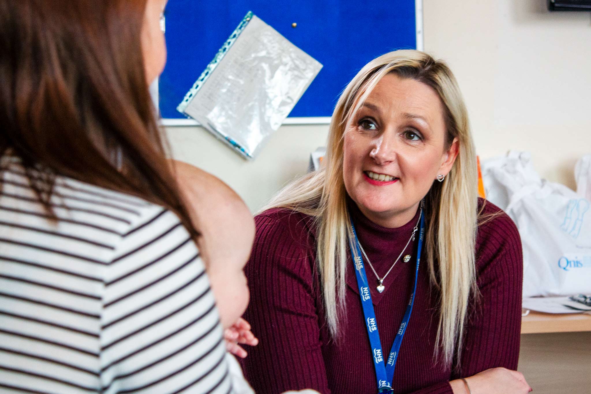 A smiling person with long, blonder hair, wearing a maroon sweater and a blue NHS lanyard, sits at a desk smiling warmly at a person holding a baby. The person facing them wears a striped shirt. Behind them, a blue noticeboard with a plastic folder on it, and bags of materials are visible.