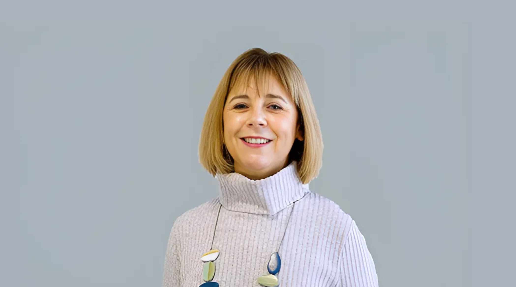 A person with straight, chin-length hair is smiling and facing the camera. They are wearing a light-coloured, ribbed turtleneck jumper and a statement necklace with large, colourful beads. The background is a plain, soft grey.