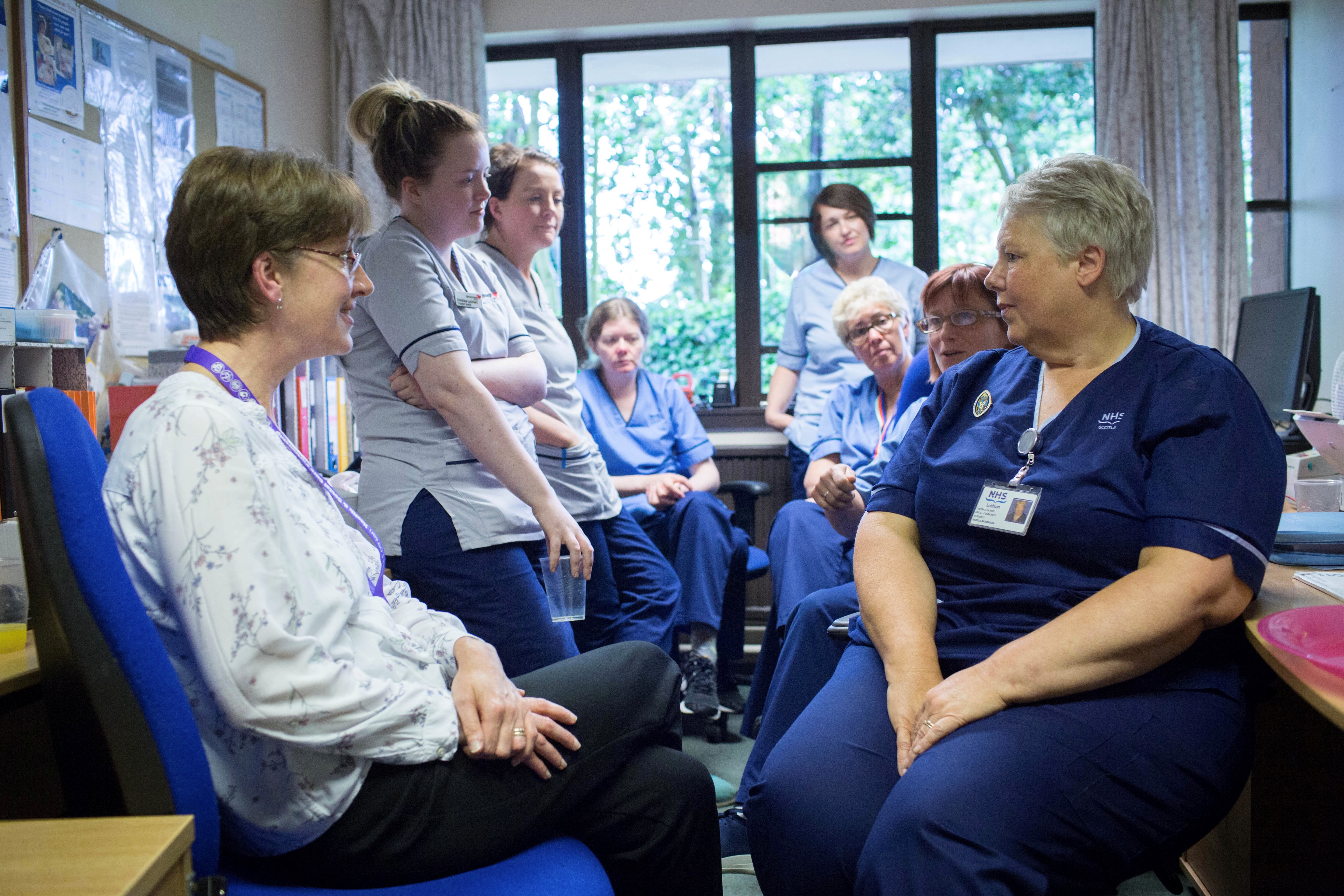 A group of people in an office, some seated and some standing, engaged in conversation. Most are wearing NHS uniforms in shades of blue and grey. In the background is a window showing greenery outside.