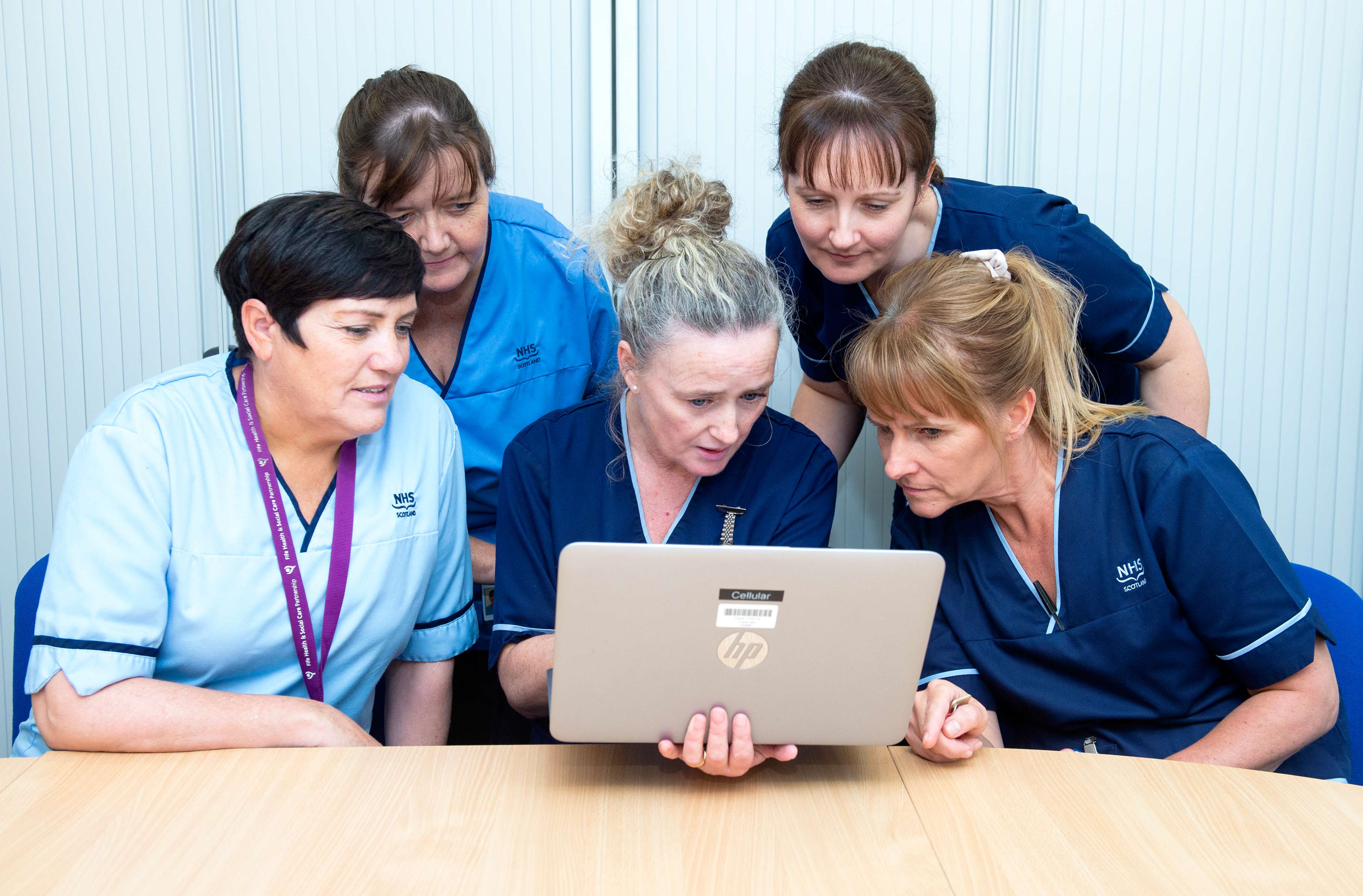 Five people wearing NHS Scotland uniforms gathered closely around a laptop, intently looking at the screen. They are seated at a wooden table in a bright indoor setting with a light-coloured background.