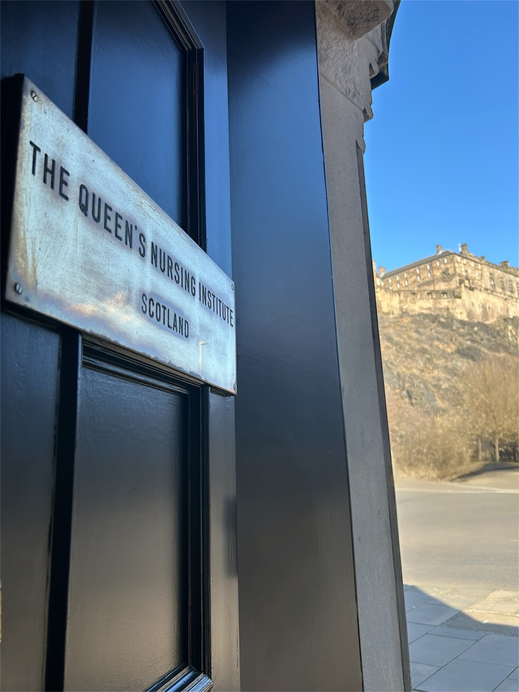 A close-up of a black door with a metal plaque reading "The Queen's Nursing Institute Scotland." In the background, across a quiet road, Edinburgh Castle is visible atop a rocky hill under a clear blue sky.