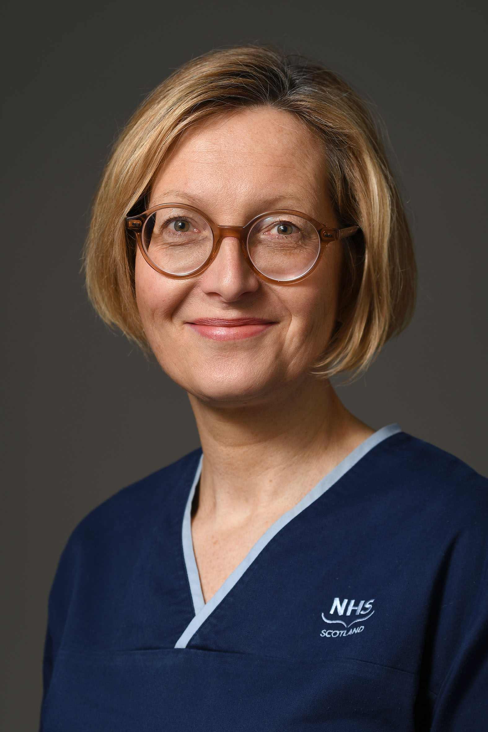 A person with short, light brown hair and round glasses is smiling slightly. They are wearing a navy blue uniform with light blue trim and an embroidered logo that reads 'NHS Scotland.' The background is plain and neutral.