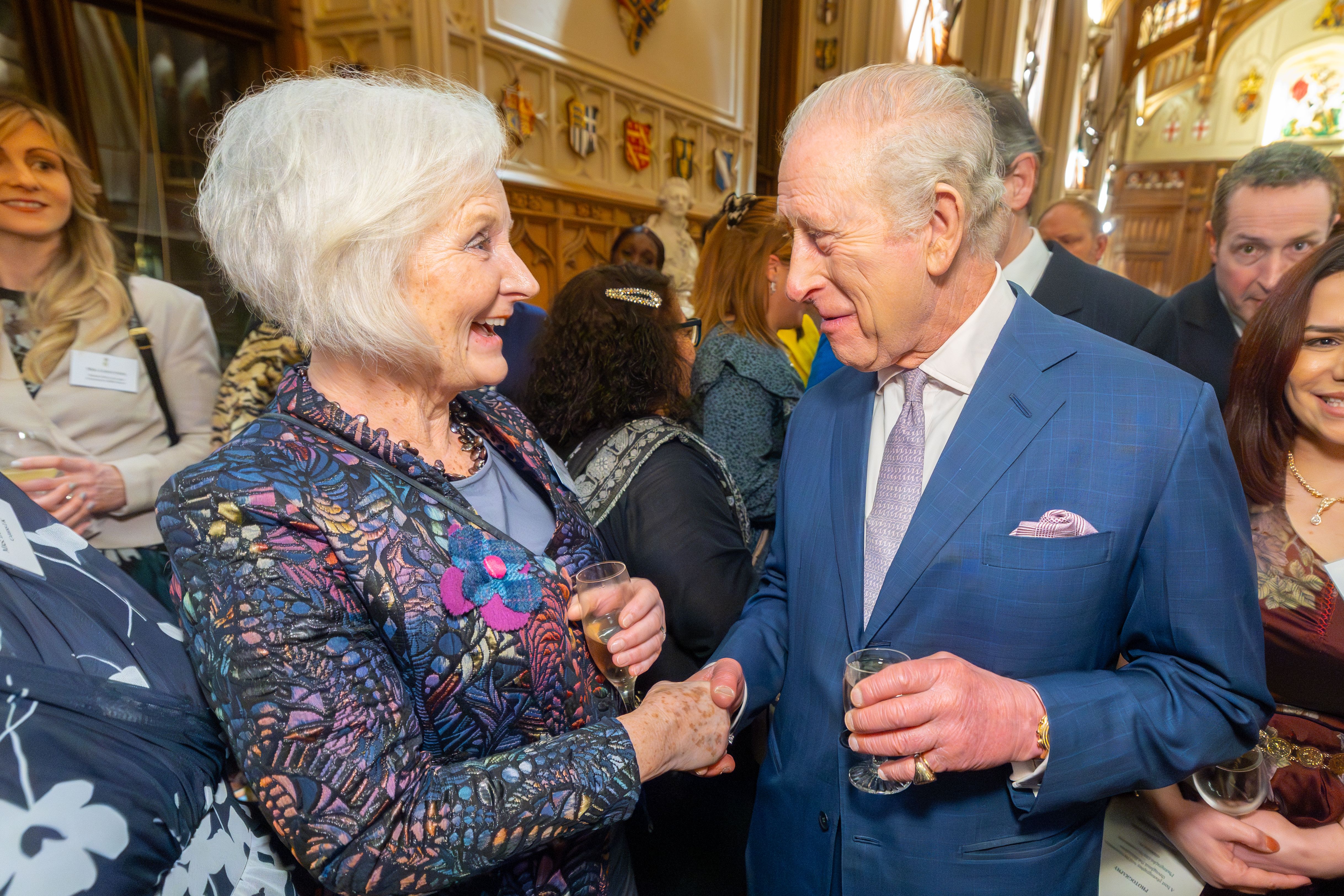 Two smiling people with white hair, each wearing a blue jacket and have a glass in their hand. Other people are visible in the background of a formal setting.