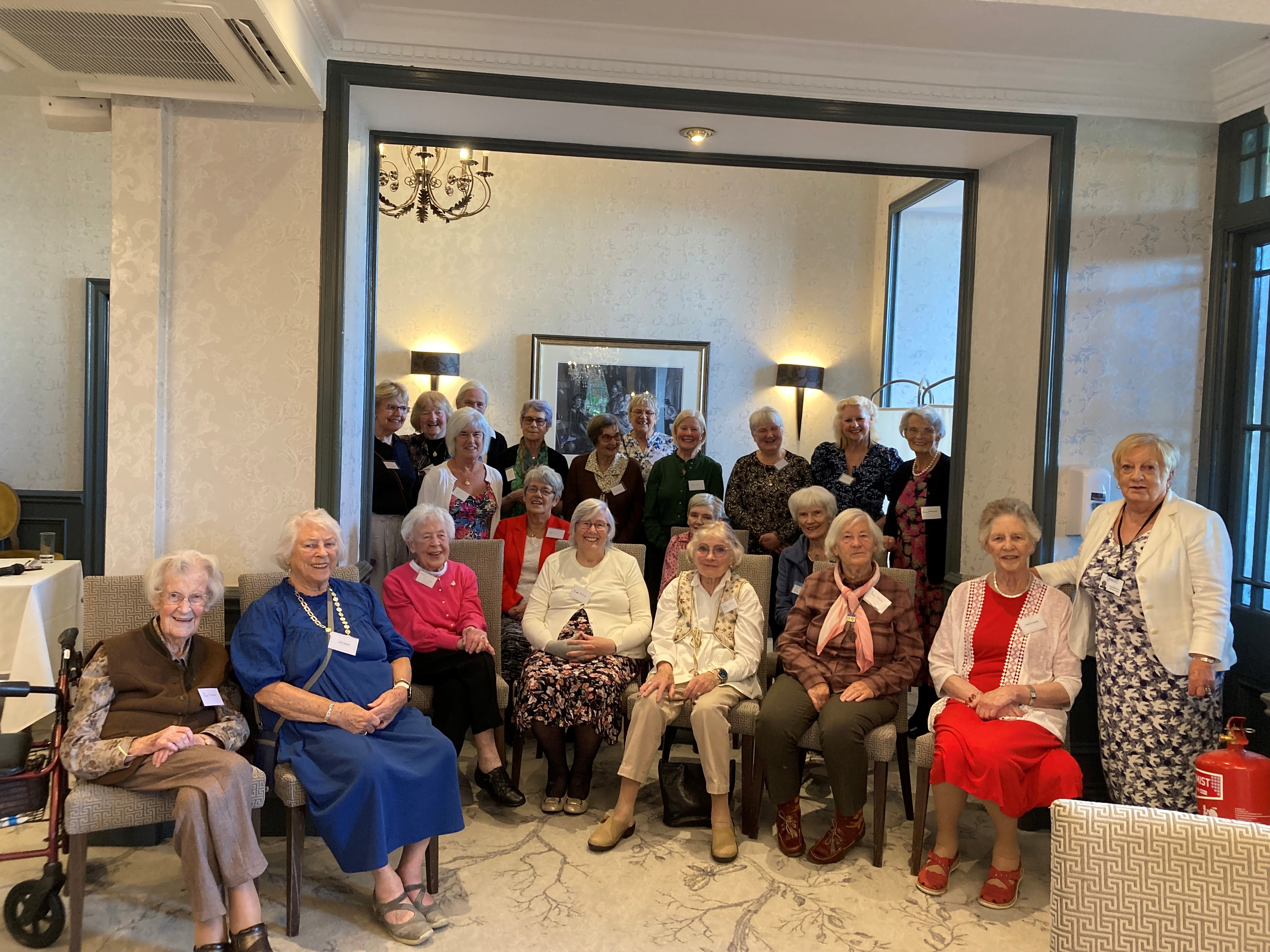 A colour image of a group of older adults smiling and seated and standing together in a large room. Some of the group stand in a large doorway or arch. There are lamps and a picture on the rear wall and a chandelier visible.