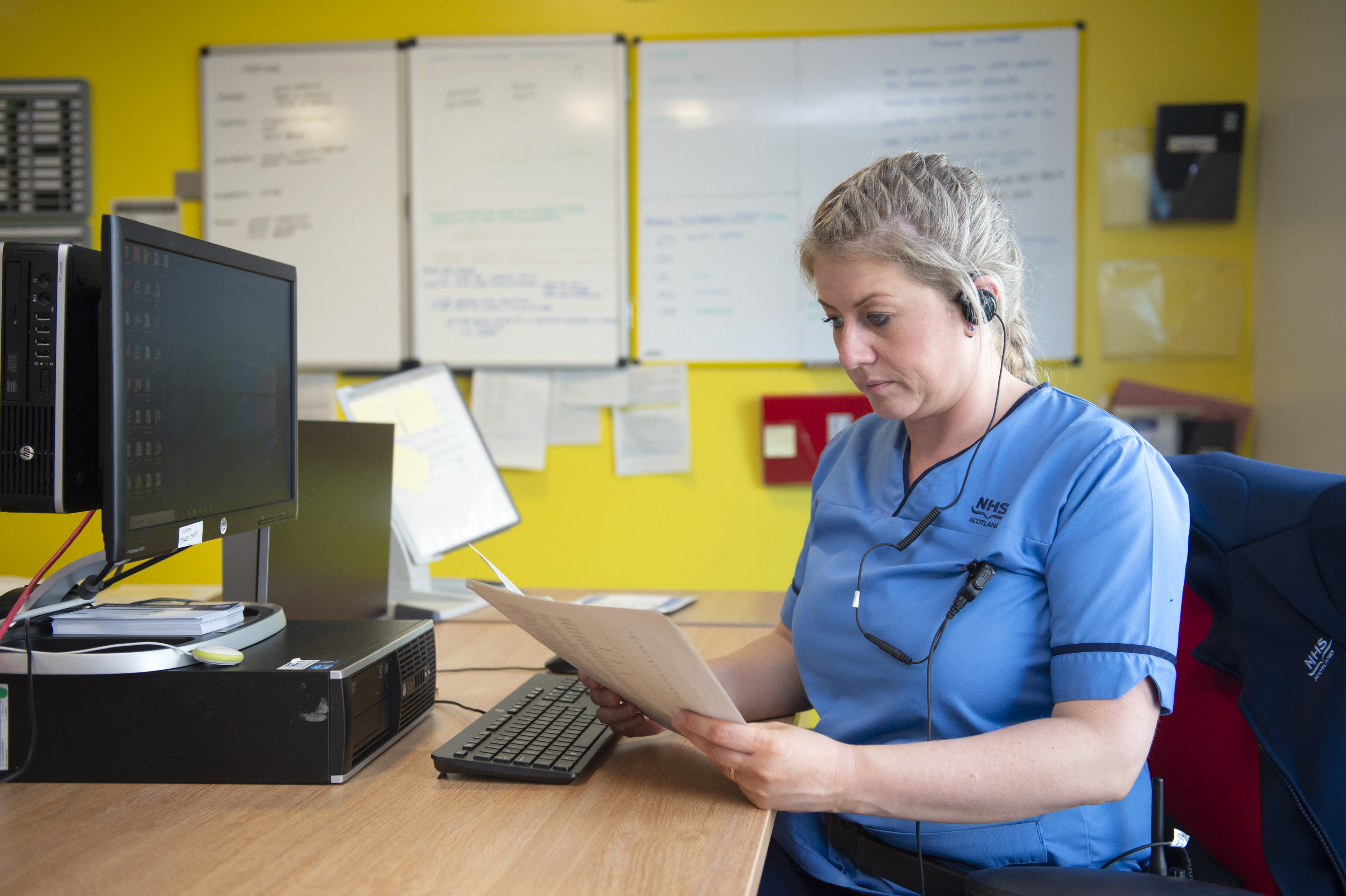 A person wearing a blue short sleeve nurse’s top with NHS on it sits at a desk holding a stack of white papers which they're looking at. They're wearing a black headset and have blonde hair in a French plait. On the desk is a computer, and in the background a yellow wall with three white noticeboards on it that all have writing on them.