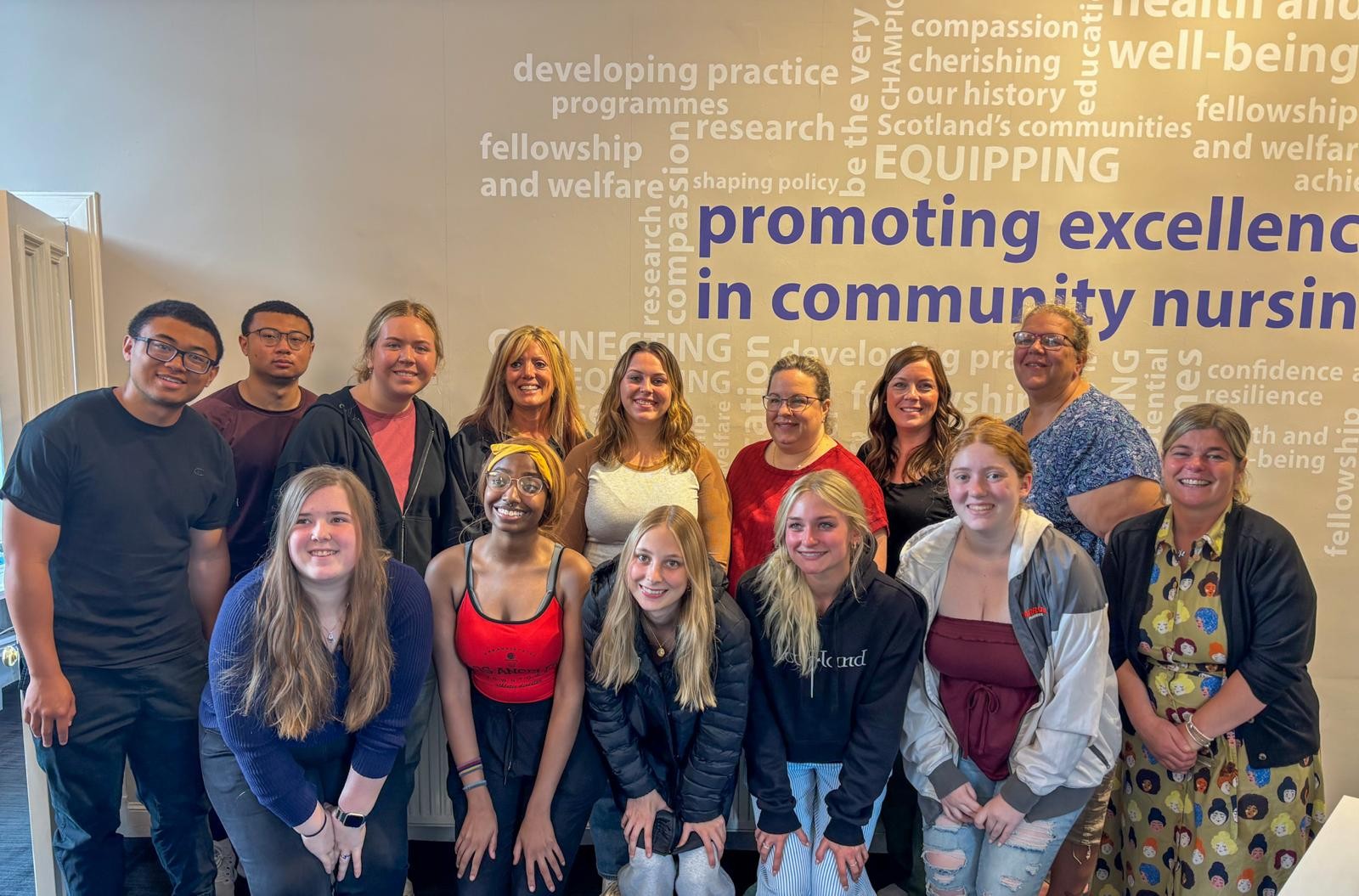 A group of fourteen people pose indoors in two rows, smiling at the camera. The people in the front row crouch slightly, while the back row stands. Behind them, a wall displays large text phrases in various fonts and orientations, including 'promoting excellence in community nursing,' along with words related to health, compassion, and research. The group is casually dressed, with varied clothing styles and colours, in a brightly lit room.