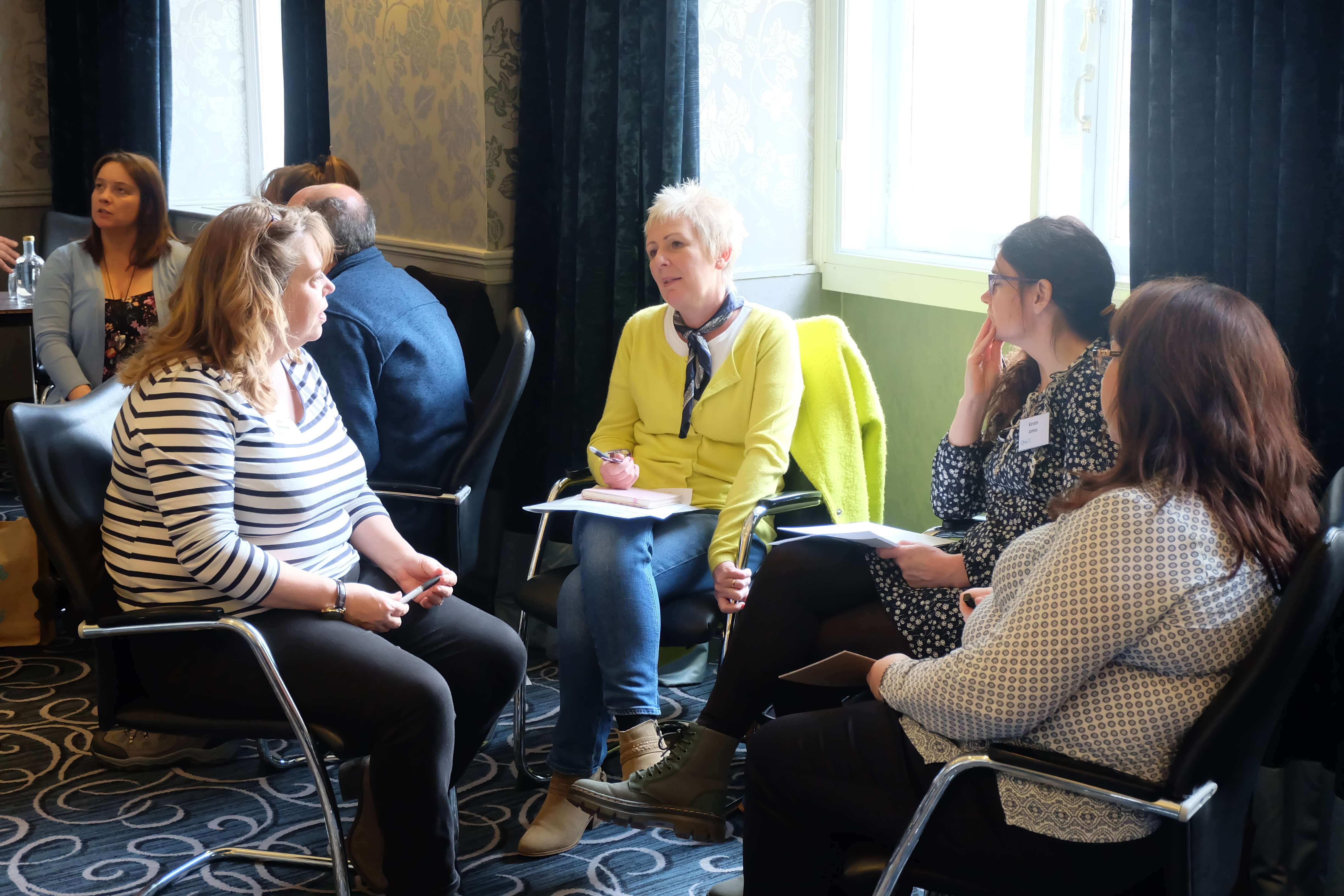 A small group of adults seated in a semi-circle during a discussion at a workshop or meeting. One person in a yellow sweater is speaking while others listen, with notebooks and pens in hand. The setting is a bright room with patterned carpet, cream wallpaper, and large windows letting in natural light.’