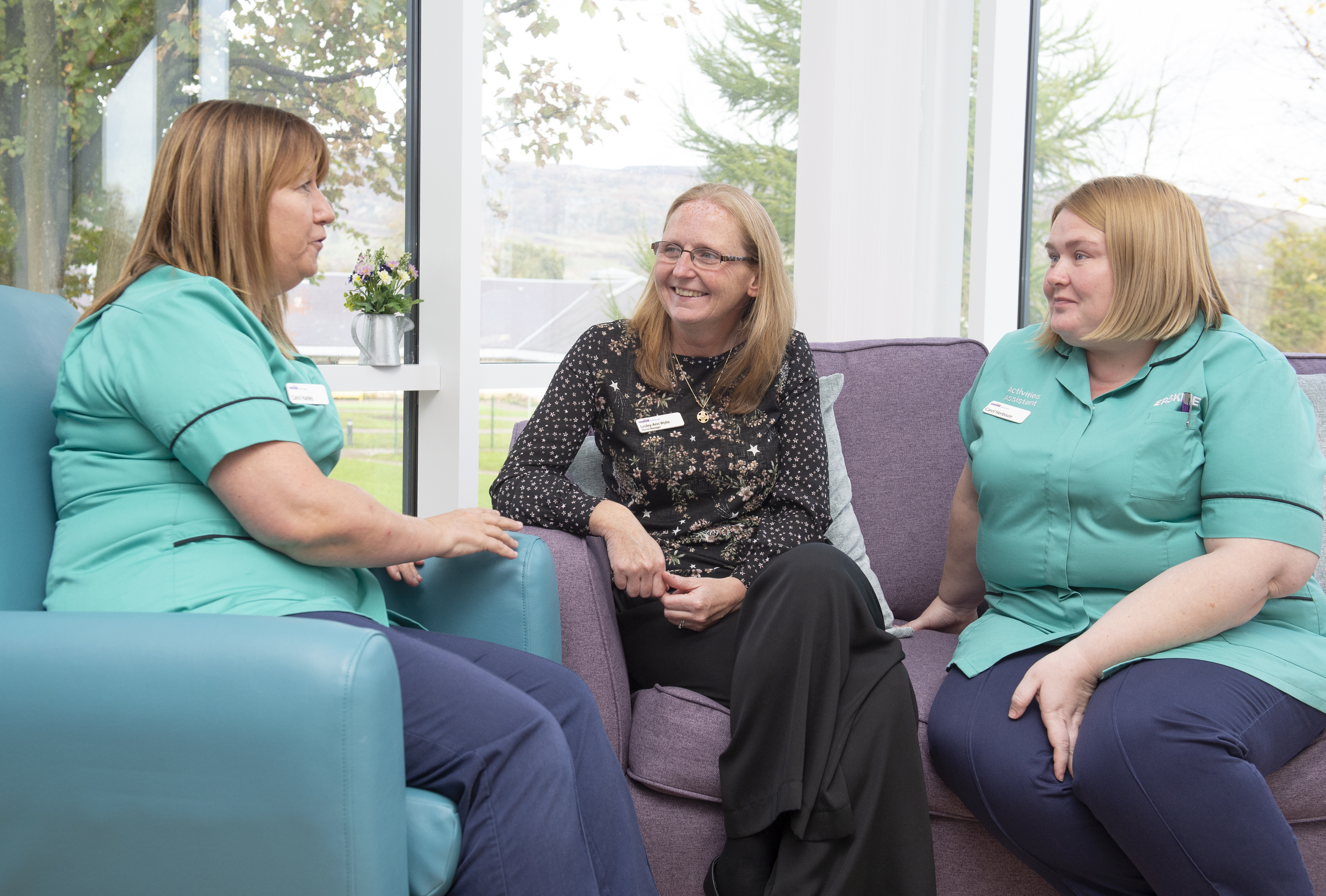A group of three people sit on a purple sofa and a turquoise chair. Two are smiling and look to be listening to the third person. The person on the left has mid brown hair past her shoulders is wearing a green nurses top with a white badge on it and blue trousers. The person in the middle has blonde hair below their shoulders and is wearing glasses, a black shirt with a white pattern of dots and flowers. 