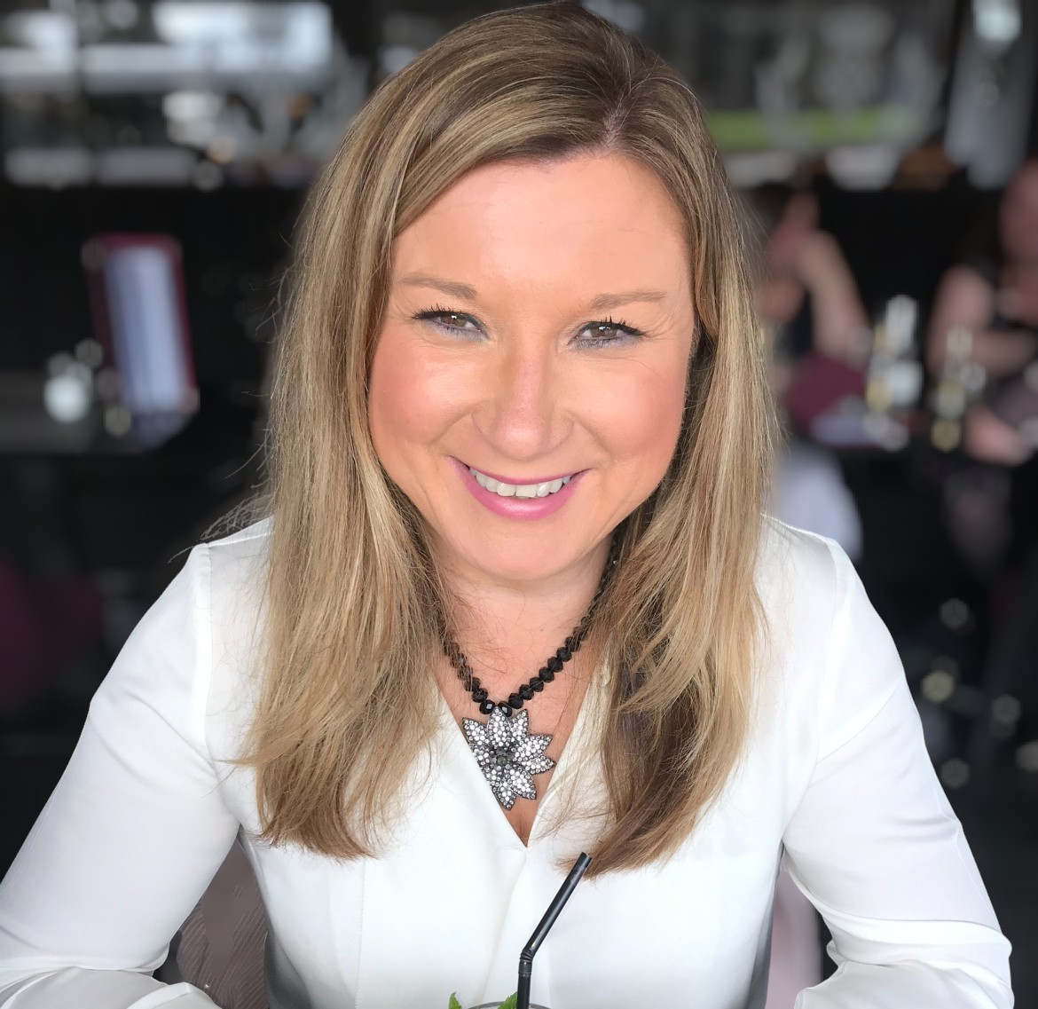 A person with long, straight, light brown hair is smiling at the camera while seated in a restaurant. They are wearing a white top and a statement necklace with a large, flower-shaped pendant. In the background, other diners are visible, and there is a window with etched decorative thistle patterns. The lighting is bright and natural.