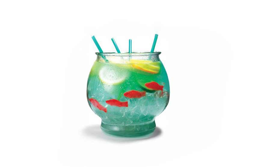 A large fishbowl-style cocktail filled with blue-green liquid, ice, lemon and lime slices, and several red gummy fish sweets. Four teal straws stick out of the top of the bowl.