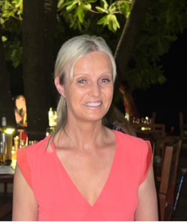 A person with light blonde hair, tied back, is smiling while standing outdoors at night. They are wearing a coral-coloured V-neck top. Behind them are wooden tables, chairs, and dimly lit dinner settings, with trees and leaves visible above.