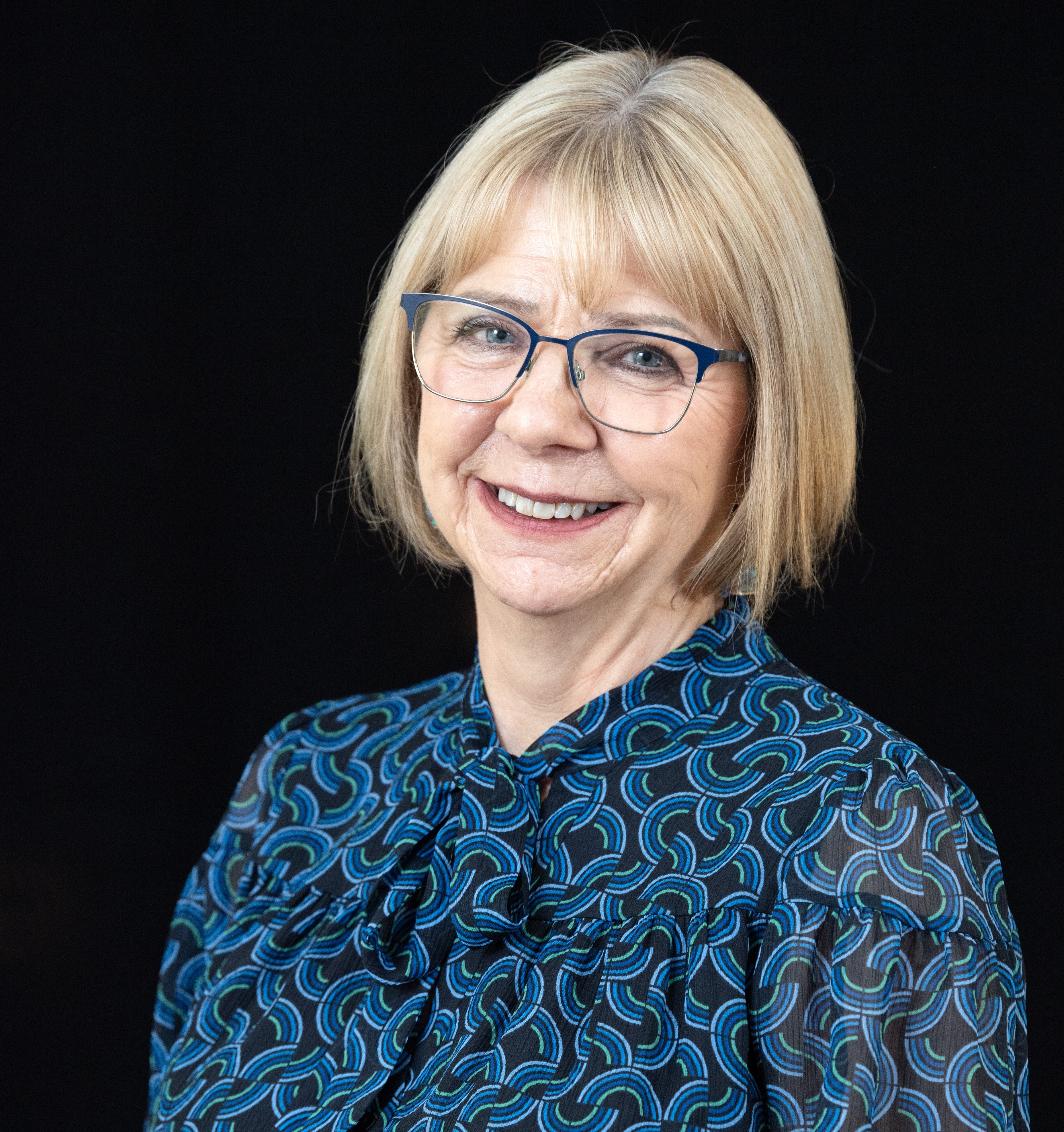 A person with short, light-coloured hair and glasses is smiling at the camera. They are wearing a patterned blouse with blue and green curved lines on a dark background. The background is solid black