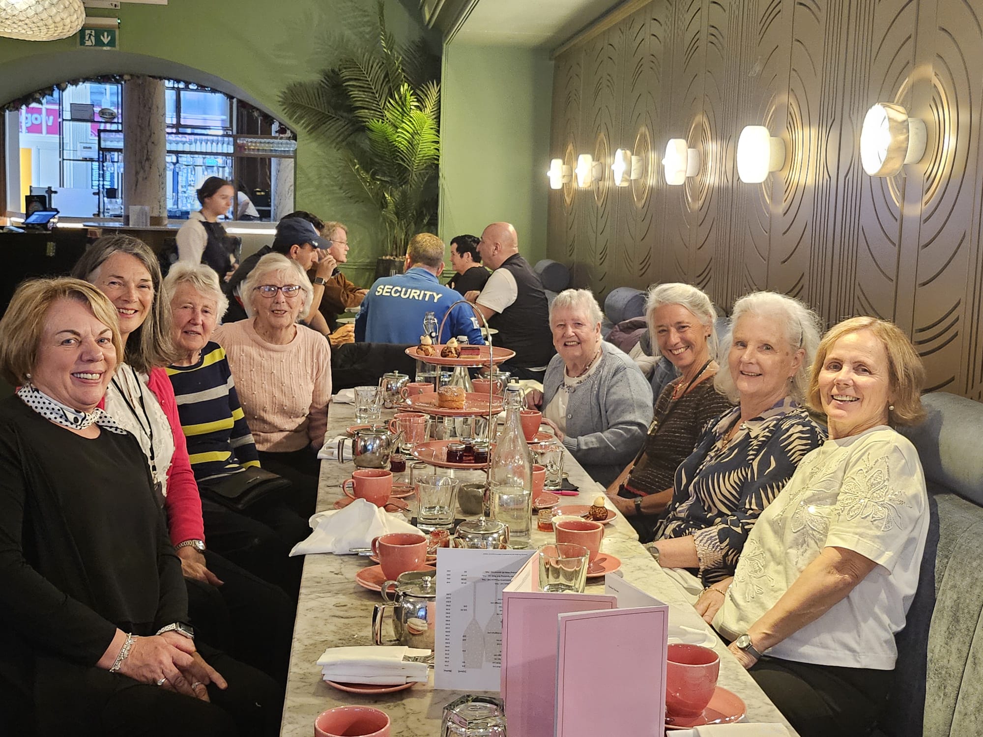 A colour image of a group of older adults smiling and seated on each side of a long table. There are menus and pink cups and saucers on the table and a row of six spotlights on the wall. A plant and a large window are in the background.