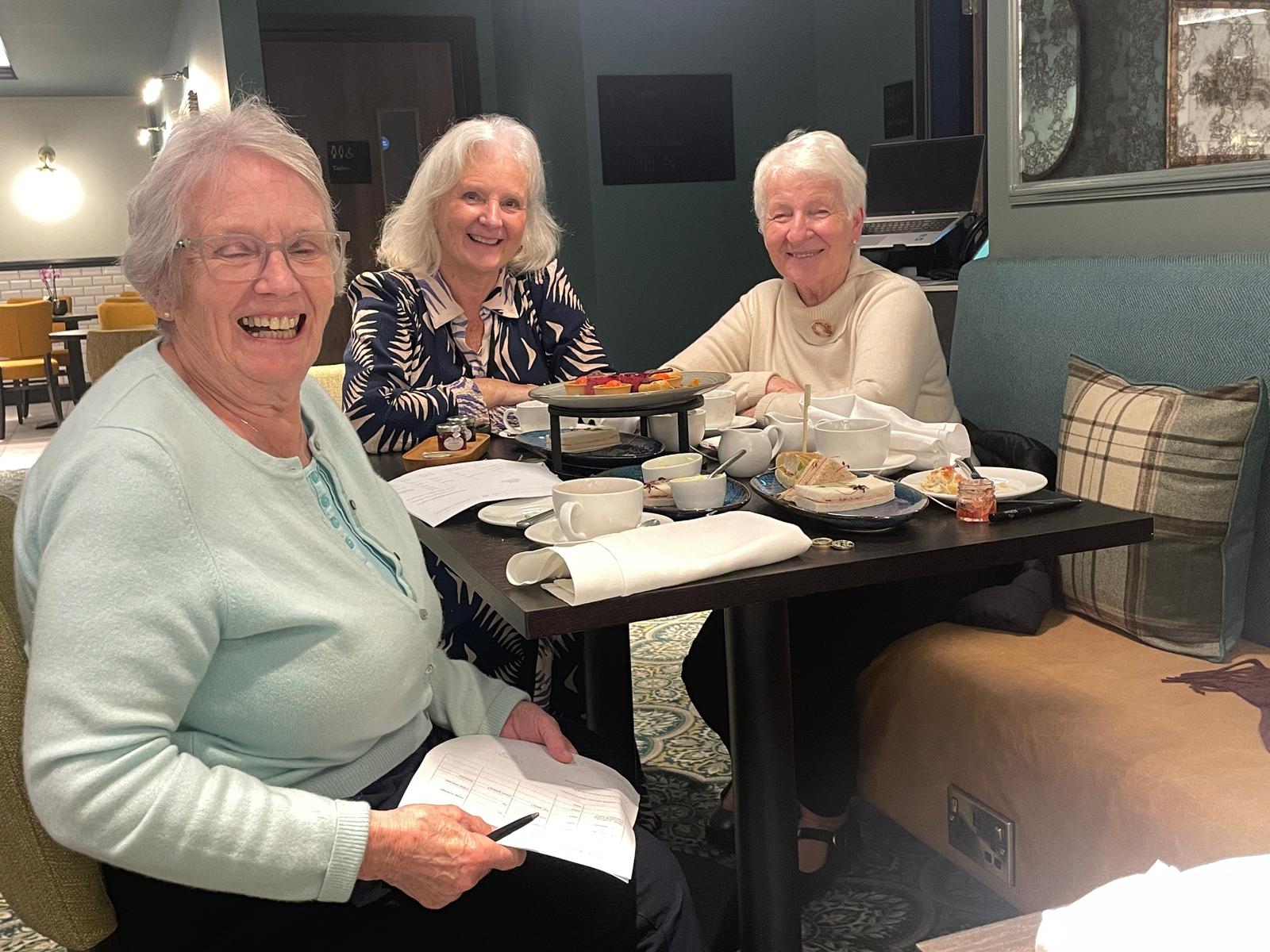 A colour image of three older adults smiling and seated around a dark wood table together.