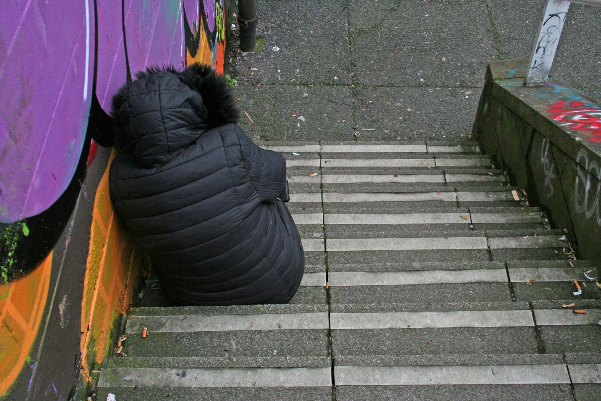 A person wearing a black puffer jacket with a fur-lined hood sits alone on a concrete stairway, leaning against a graffiti-covered wall. The scene appears urban, with litter scattered around and worn steps indicating frequent use.