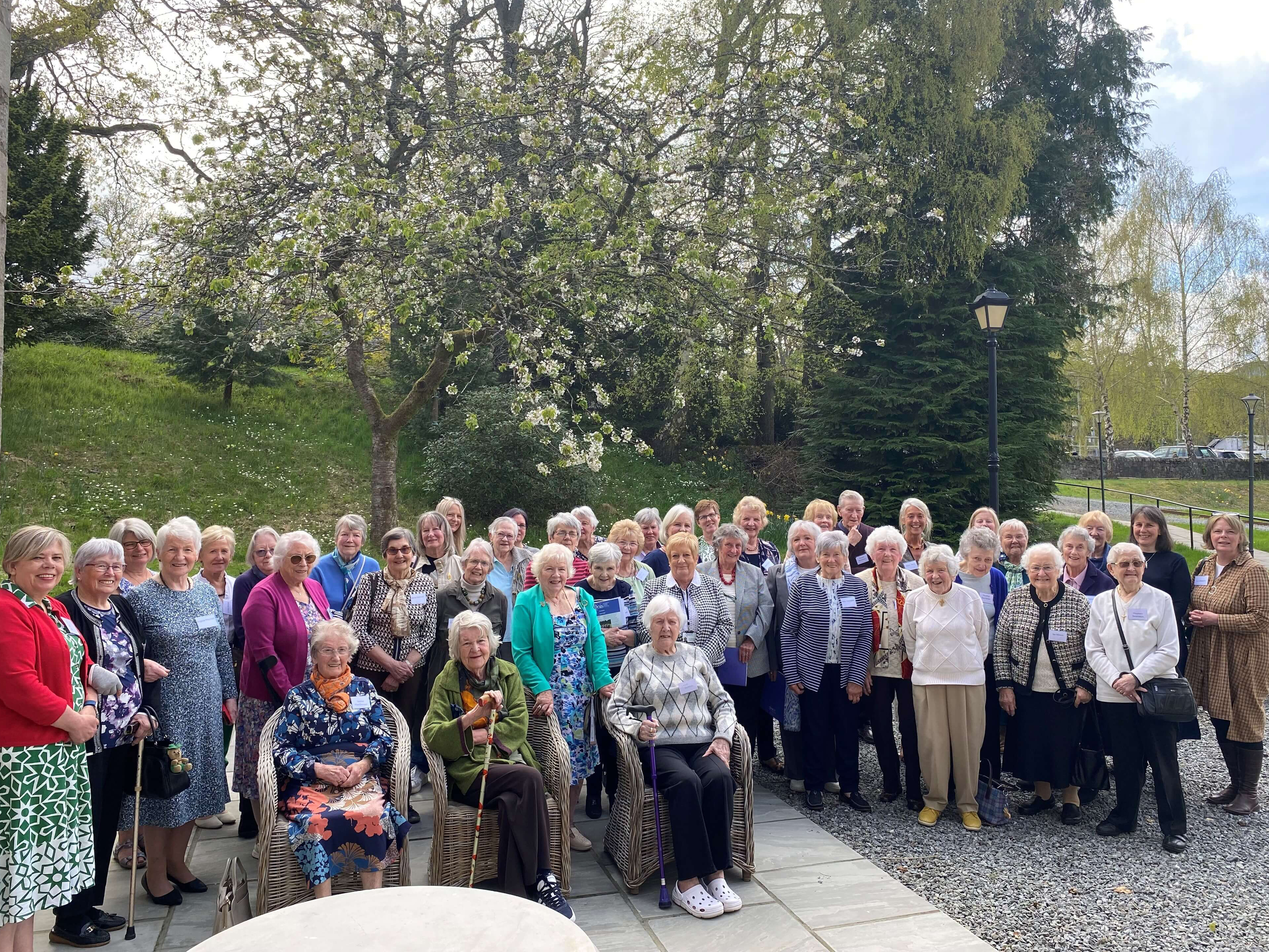 A large group of older adults gathered outdoors, standing and seated in front of blooming trees and greenery. Many are smiling at the camera, dressed in a variety of colourful and patterned clothing. The setting is a paved area with wicker chairs under a bright, partly cloudy sky.