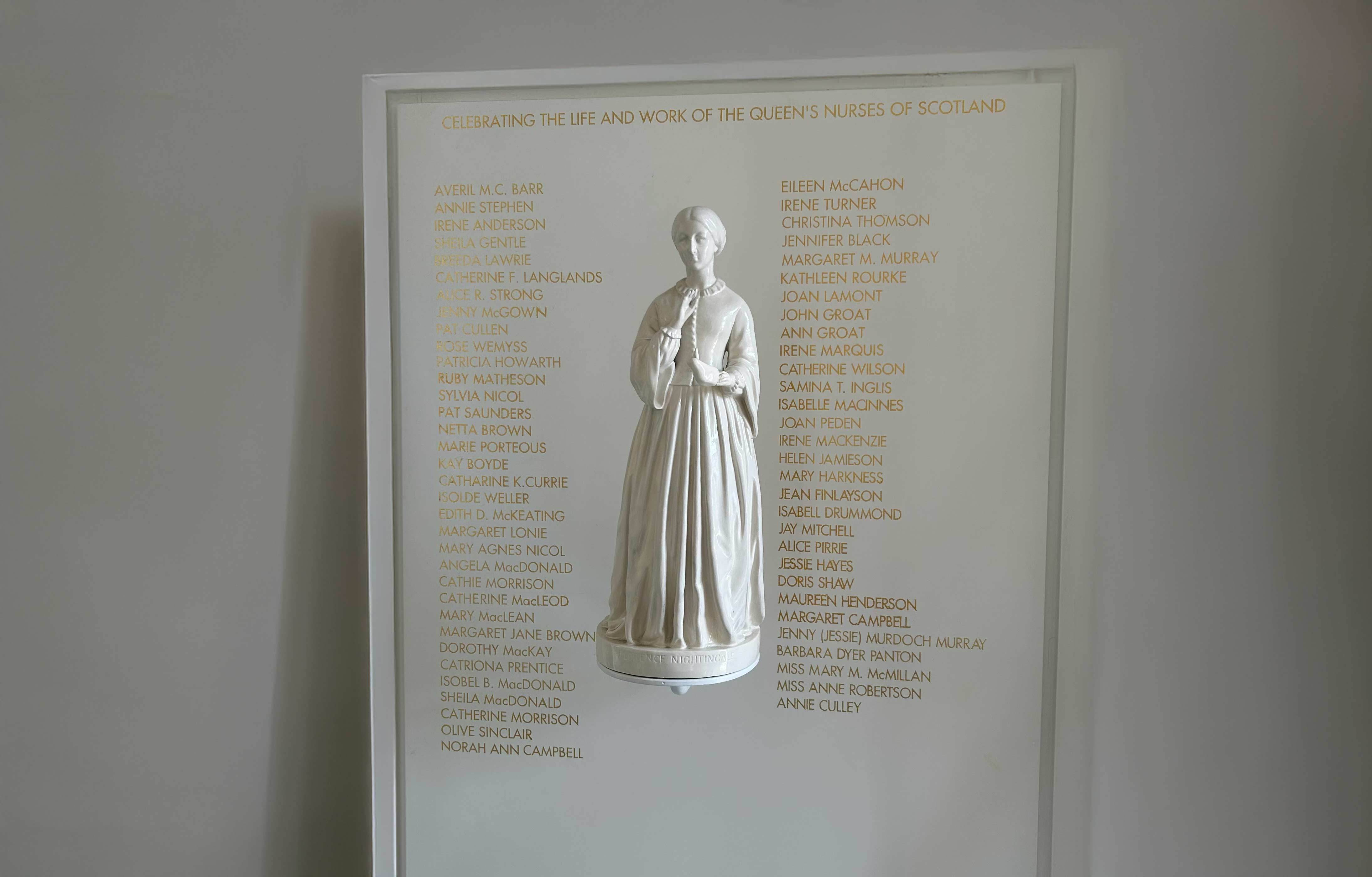 A commemorative wall display honouring the Queen’s Nurses of Scotland, titled “Celebrating the Life and Work of the Queen’s Nurses of Scotland.” A central white sculpture of a historical nursing figure, Florence Nightingale, is surrounded by two columns of names written in gold lettering.