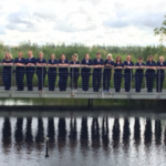 A row of people wearing nurse uniforms stand on a bridge. There is a big blue sky in the background. The people are reflected in the water below them