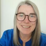 A colour image of a smiling person with blonde hair below their shoulders. They wear glasses and a blue top