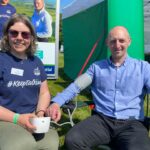 Two smiling people sitting outside in the sunshine. The person on the left wears sunglasses, green trousers, and a dark blue T-shirt with white text that says #KeepTalking. The person on the right wears black trousers and a light blue long-sleeved shirt. They have a blood pressure monitor around one arm